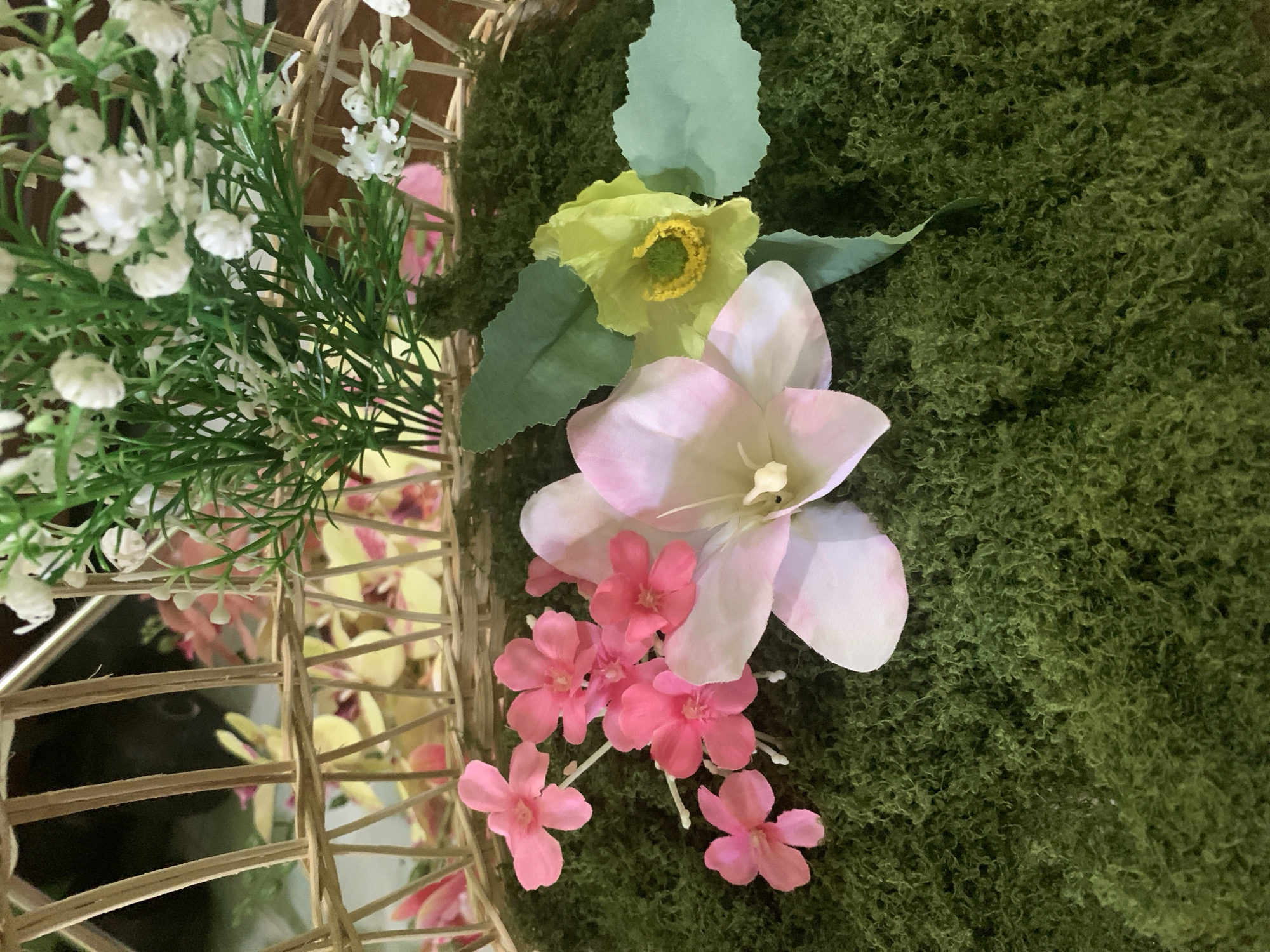 This is a close-up, eye-level, indoor shot of an artificial flower arrangement in a woven basket, with no visible people. The basket is filled with green moss, on which several artificial flowers are placed. The flowers include a large pink and white orchid-like bloom, a cluster of small pink flowers, and a single yellow flower with a green center. Behind the moss, the base of the basket is visible, with more artificial flowers, predominantly yellow and pink, peeking through the woven structure. In the upper portion of the image, a bundle of small white artificial flowers and green foliage is visible, also within the basket. The background is dark and out of focus, suggesting an indoor setting, possibly a display or decoration. There is no indication of the location being in Kuala Pembuang, Indonesia, or any city.
