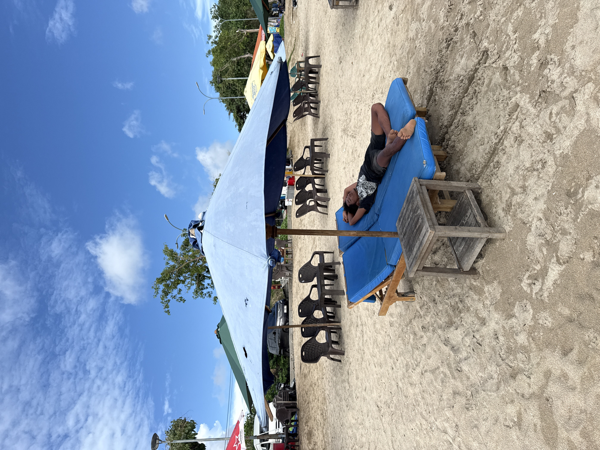 The image depicts a bright, sunny scene at a sandy beachfront location, likely in Kuta, Indonesia. In the foreground, an individual, appearing to be an adult male, is comfortably reclining on a blue mattress atop a wooden beach lounger. He wears a dark t-shirt and shorts, with his bare feet visible, and looks casually towards the camera, seemingly at ease.
A large, light blue beach umbrella dominates the left side of the frame, providing shade over the seating area. Under and around this umbrella, as well as extending into the midground, are numerous dark plastic chairs, some stacked and others arranged for use. Further back, more colorful beach umbrellas (yellow and green) are visible, along with lush green tropical trees and foliage, hinting at a vibrant natural setting. In the distant background, elements suggesting a road or pathway with parked vehicles and street lights can be discerned.
The sky is a clear, brilliant blue with scattered, fluffy white clouds, indicating excellent, sunny weather during daytime, possibly morning or early afternoon. The ground is entirely covered in light-colored sand, showing subtle textures and footprints.
The overall atmosphere suggests leisure and relaxation typical of a popular beach destination. A notable detail in the background includes a red banner or umbrella displaying partial text, including "BITANG" and possibly "COLA," which are common brands in Indonesia, reinforcing the specified location context. The scene captures a peaceful moment of rest amidst typical beach amenities.