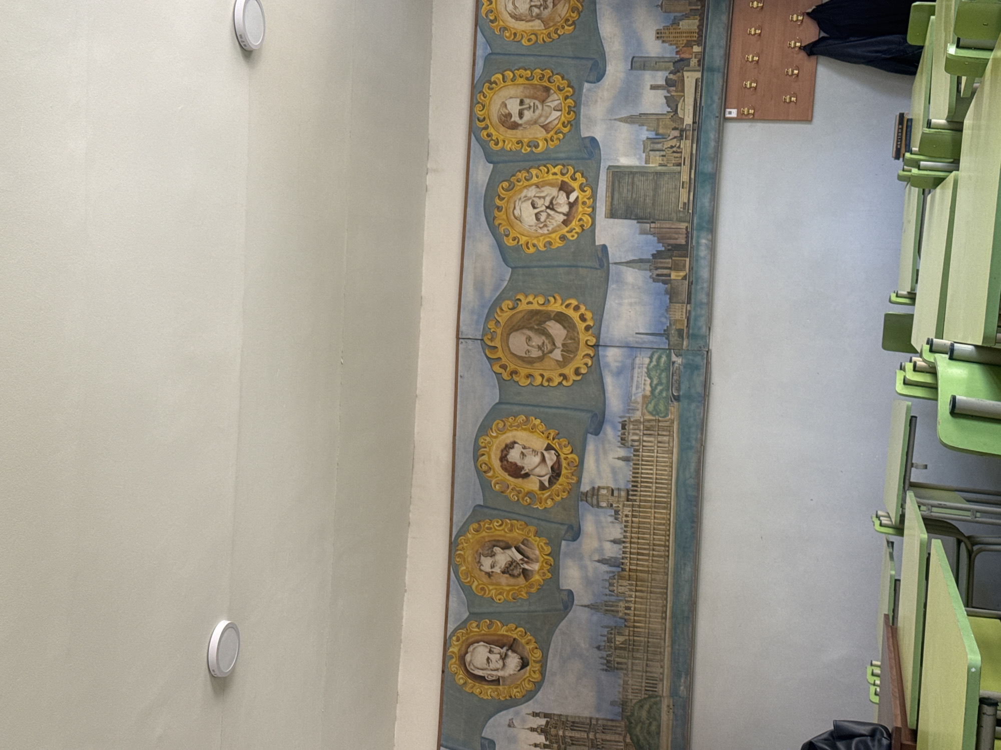 The image depicts a classroom or educational space, characterized by a detailed mural on one wall and light green and wooden desks arranged along the opposite wall. The room is brightly lit by unseen sources, with light beige walls.
The prominent feature is a large, vertically oriented mural composed of multiple painted panels. This mural showcases a series of historical or influential figures, each depicted in a golden, ornate oval frame set against a blue scroll-like background. From top to bottom, the portraits include a man with a mustache, a woman with dark hair, a man with a beard and mustache, a man with a full beard, a man with glasses and a white beard, a man with a goatee reminiscent of William Shakespeare, and a man with a prominent white beard and bald head. Interspersed with these portraits are painted cityscapes. One section features a modern city skyline, possibly including the United Nations building, while another clearly depicts the Houses of Parliament and Big Ben along the River Thames in London.
On the right side of the image, multiple light green desks with light wooden tops are visible, some stacked, suggesting a classroom setup. A book with the word "ENGLISH" written on its spine is partially visible among the desks. Above the desks, a wooden coat rack with several brass hooks is mounted on the wall, with a black bag or jacket hanging from one hook. The left wall is plain light beige, featuring two circular white devices, possibly smoke detectors or lights, mounted at different heights.