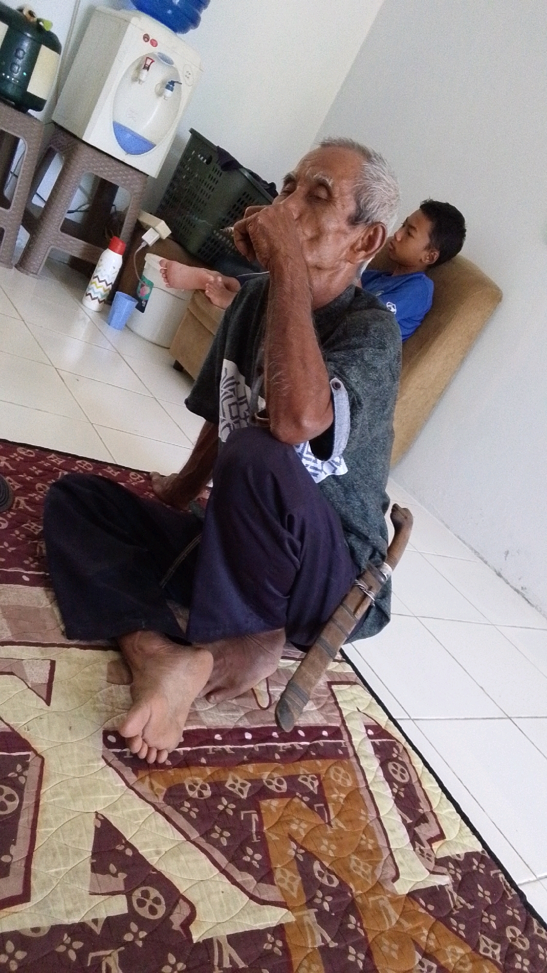 An elderly man with graying hair and closed eyes is sitting on a patterned rug, with his hand to his mouth. He is wearing a dark t-shirt and dark pants. A dagger is tucked into his waistband. Behind him, a younger boy in a blue shirt is lying down on a tan couch. In the background, a water dispenser and a laundry basket are visible. The flooring appears to be white tiles. The image seems to be taken indoors in what could be a home in Cintakarya, Indonesia, given the context of the location.