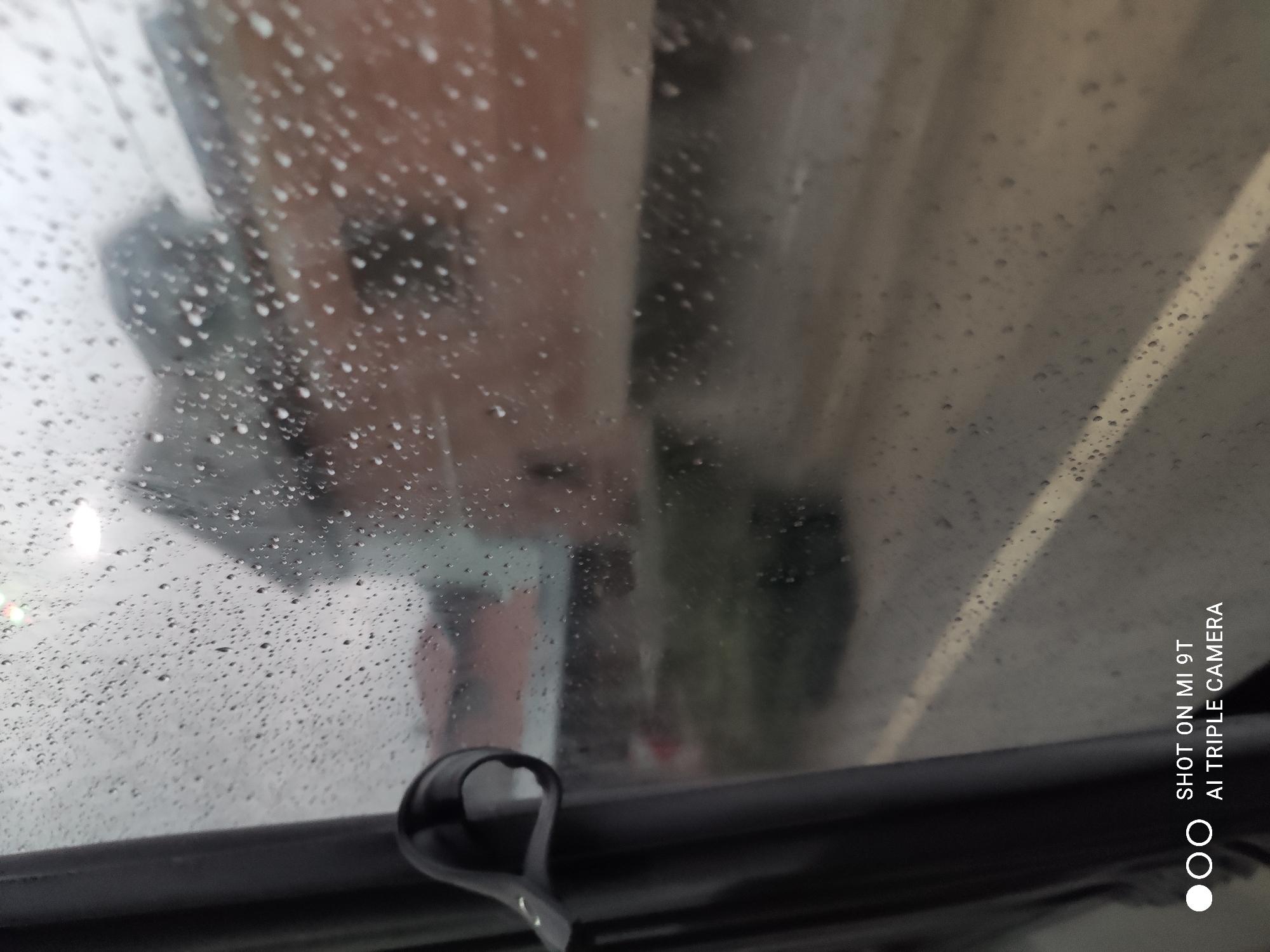 The image captures a view through a rain-streaked window in Akçaabat, Turkey. A dark grey curtain is visible on the left side of the frame, with a black fabric loop attached to it. The window is covered in numerous water droplets, distorting the view of the outside environment. Beyond the window, a blurred brick building is discernible, with hints of a red roof. A road with a white line is also visible, suggesting an urban setting. The sky appears overcast and grey, consistent with rainy weather. The bottom of the image displays text that reads "SHOT ON MI 9T AI TRIPLE CAMERA".