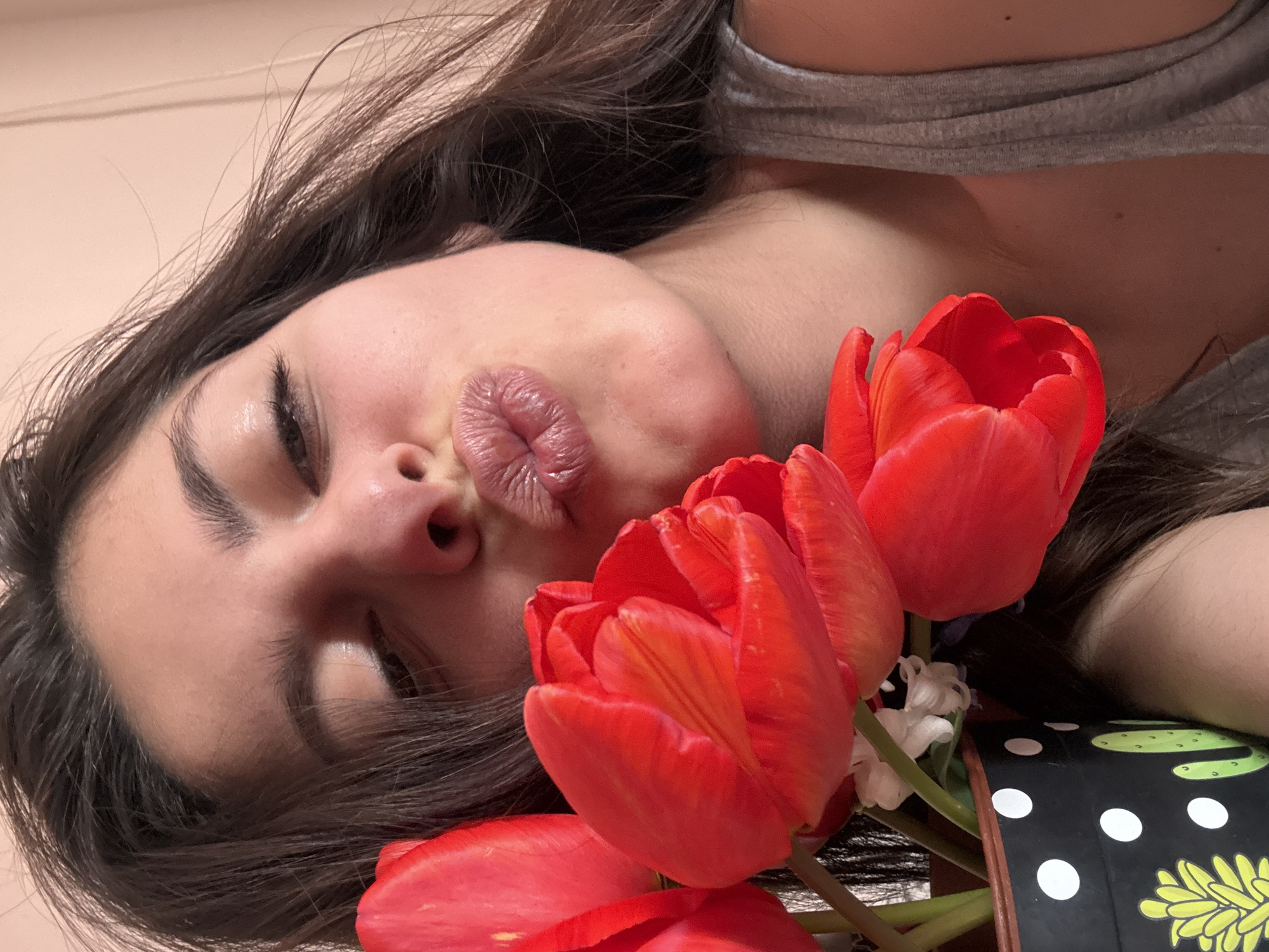 A young woman lies on her side, eyes closed, puckering her lips in a kiss. Her dark hair is partially visible. She is wearing a gray top. Next to her face are three red tulips and a small white flower in a bouquet, wrapped in a black paper with white polka dots. A green cactus design is also partially visible on the paper. The background is a pale peach color. The location is not identifiable as Plovdiv, Bulgaria based on the visual information provided.