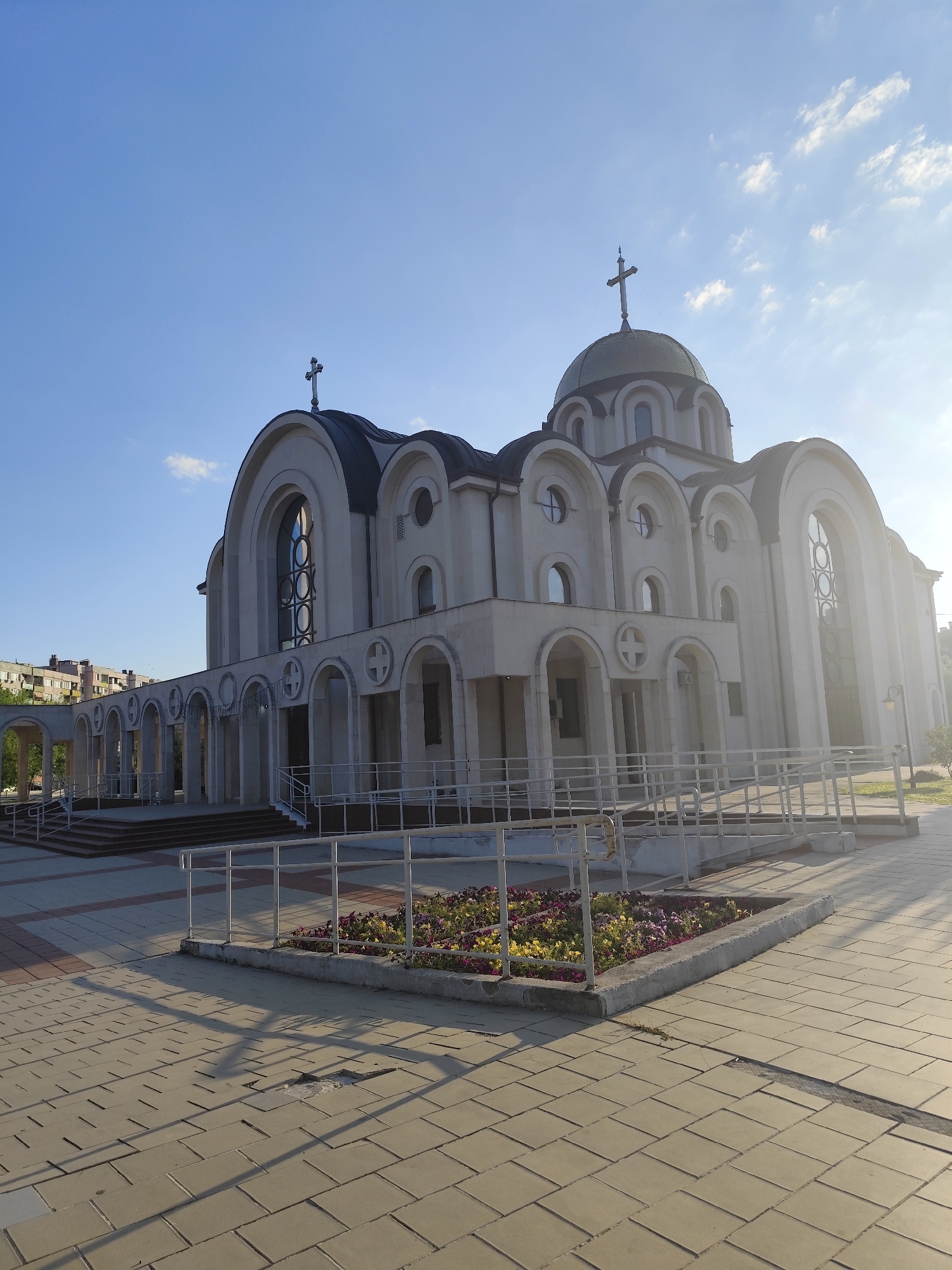 The image shows the Church of St. George the Victorious in the city of Montana, Bulgaria. The church is a modern building with a prominent dome topped with a cross, and several smaller domes and arched windows. The exterior is a light beige color, with dark grey roofing. In front of the church is a paved plaza with a flower bed containing colorful flowers. There are also ramps and stairs leading up to the entrance of the church, with metal railings. The sky is blue with scattered white clouds, and the sun appears to be setting, casting long shadows across the plaza. In the background, there are apartment buildings. No people are visible in the image.