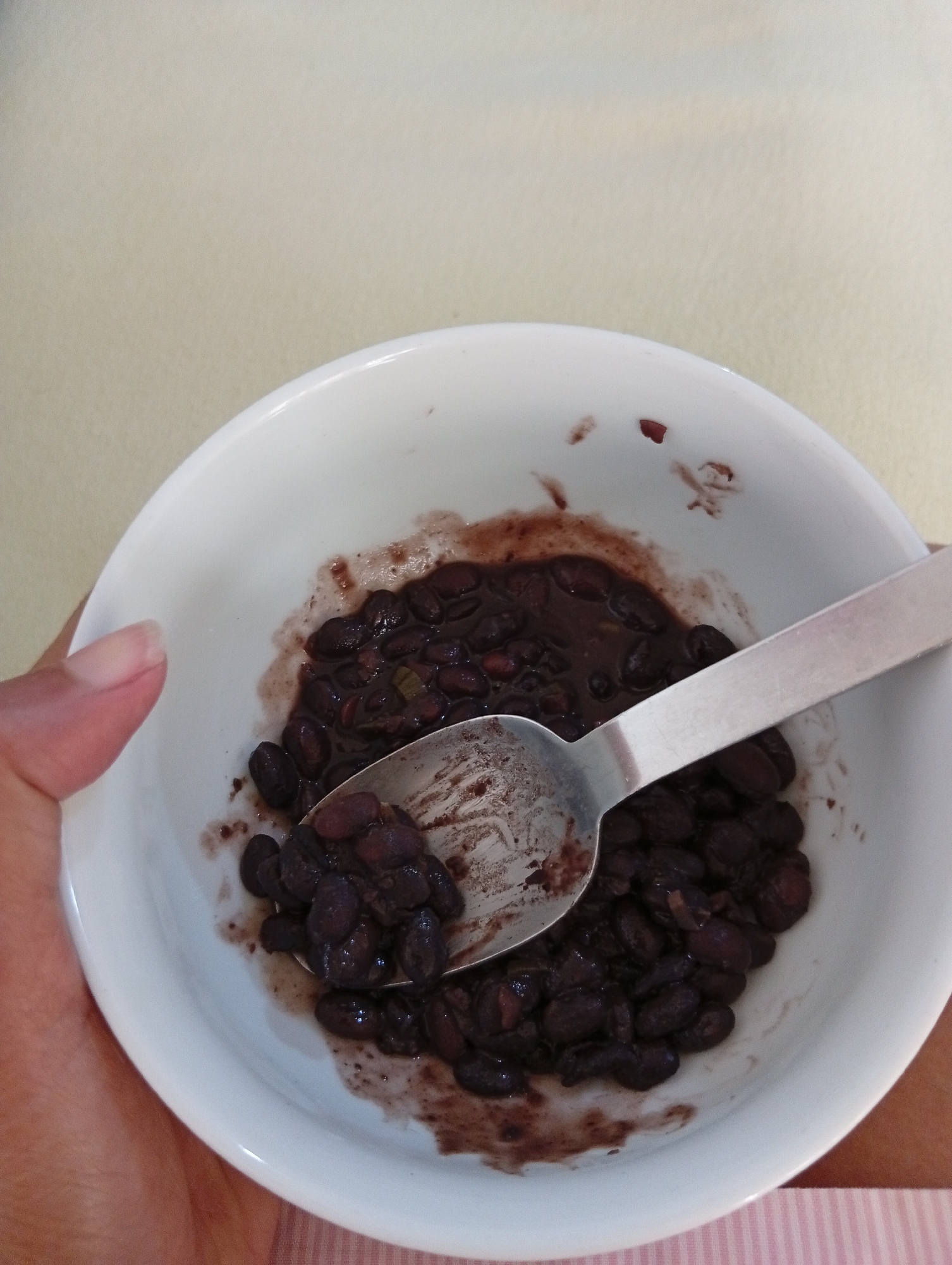 A close-up, high-angle shot shows a white ceramic bowl filled with black beans. A silver spoon rests in the bowl, scooping up a portion of the beans. The beans appear to be cooked in a dark sauce, with some visible pieces of green onion. A person's hand, with a visible fingernail, is holding the bowl from the left side of the frame. The background is a plain, light-colored surface. The image was captured in San Antonio de Los Altos, Venezuela.