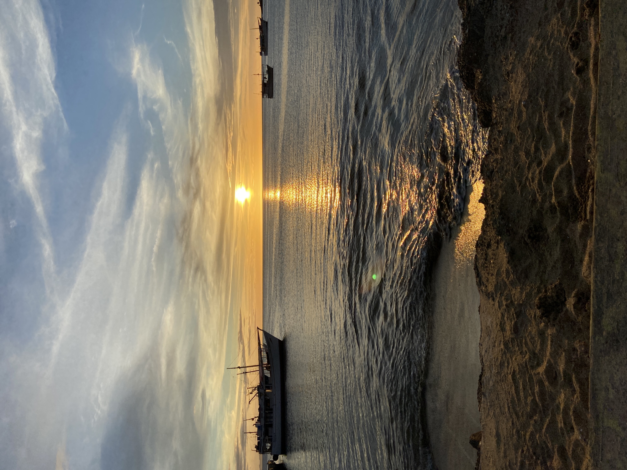 This image depicts a sunset scene in the city of Juan Griego, Venezuela. The sun is low on the horizon, casting a warm, golden light across the water and sky. The ocean is calm with gentle waves lapping at the sandy shore in the foreground. Several boats are visible on the water; a larger white boat is docked on the left side of the image, and two smaller boats are further out in the distance. The sky is a mix of blue and orange hues, with wispy clouds streaking across it. The sandy beach in the foreground has some rocks and debris scattered on it. The overall atmosphere is serene and picturesque.