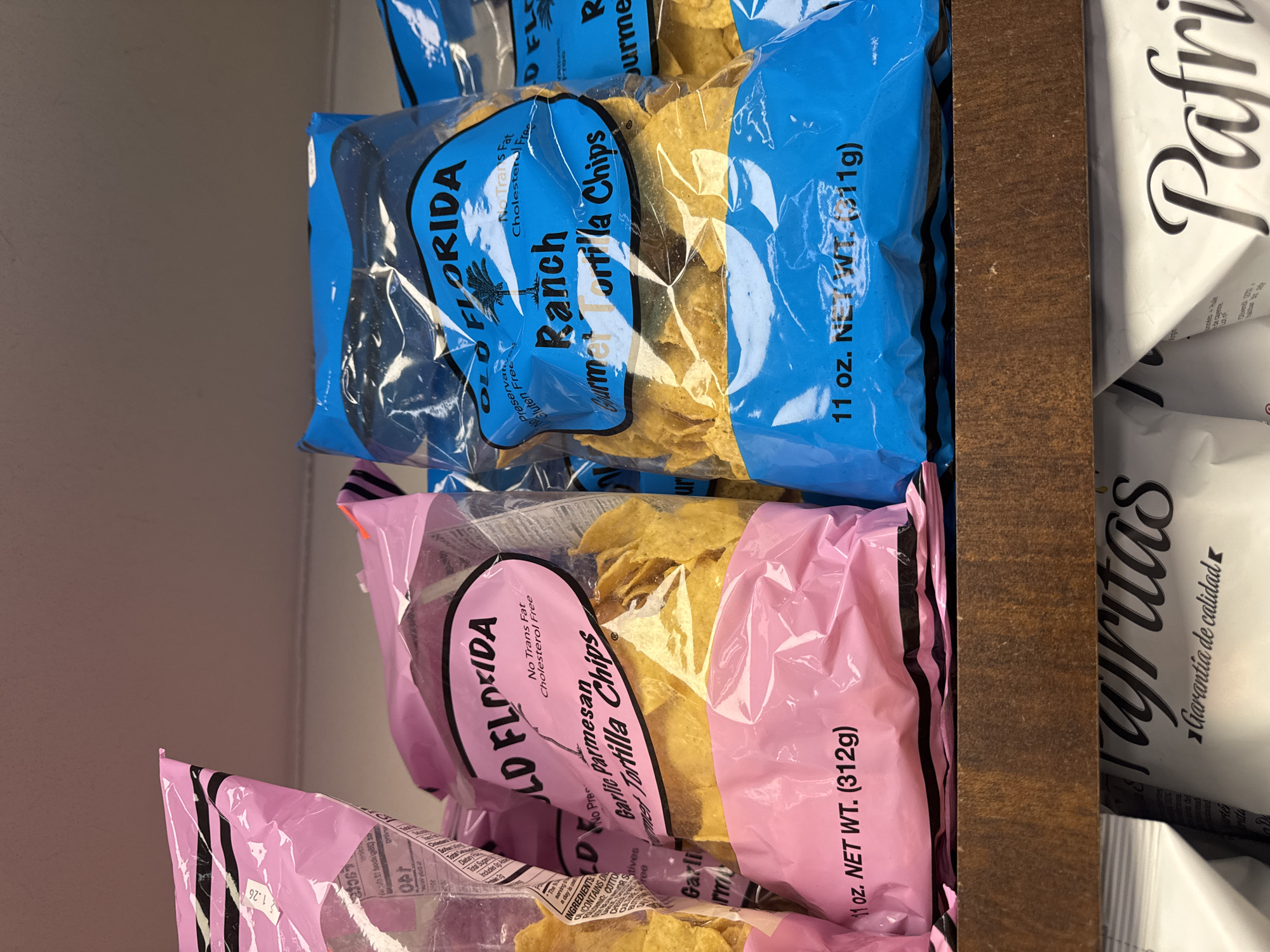 The image shows a close-up of grocery store shelf stocked with bags of "Old Florida Gourmet Tortilla Chips." The top row features blue bags labeled "Ranch" with a net weight of 11 oz (311g), while the bottom row has pink bags of "Garlic Parmesan" with a net weight of 11 oz (312g). Part of another product labeled "Tajritas" can be seen as well. The setting appears to be a typical retail environment, likely a grocery store.