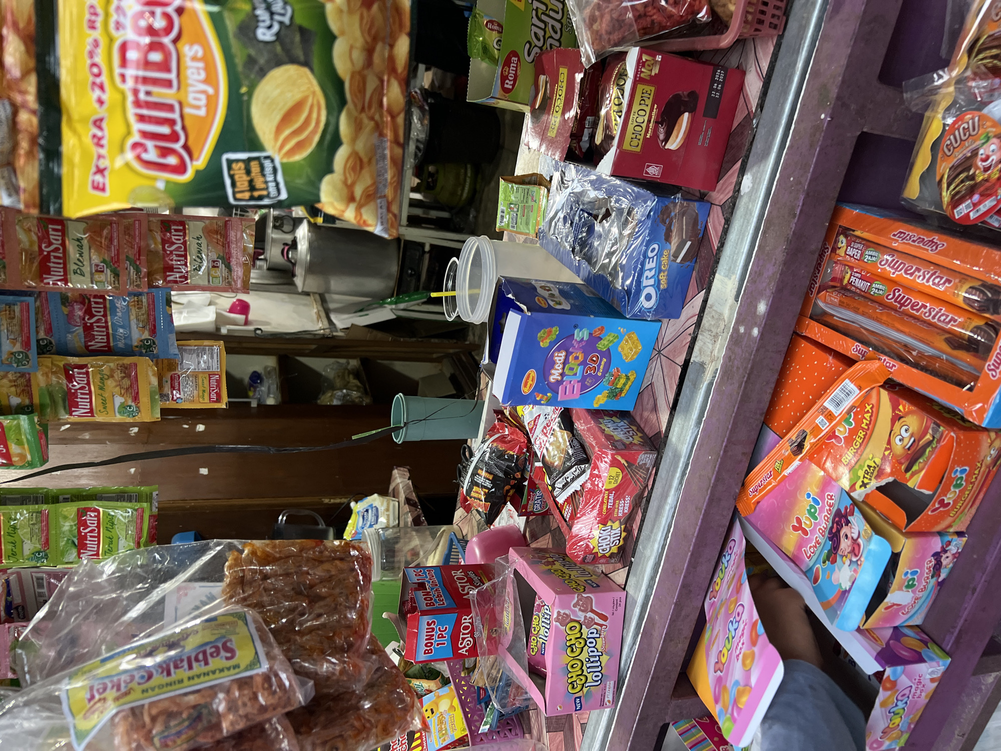 This is a medium shot of a store shelf in Gunung Putri, Indonesia, overflowing with a variety of snacks and candies. The foreground features a row of colorful boxes and packets of sweets. Prominently displayed are pink boxes of "Cho Cho Lollipop" and red boxes of "Choki Stix" and "Choco Pie" with marshmallow filling. To the left, a person's hand is partially visible reaching into a display of "Yupi" brand gummies, including "Love Burger" and "Magic Box" varieties. In the middle, blue boxes of "Oreo Soft Cake" and "Modi Blocks 3D Gummy" are stacked. Behind these, a large yellow bag of "GuriBer Layers" potato chips is the most visible item in the background, alongside other packaged snacks like "NutriSari" drinks and "Roma" crackers. The overall environment appears to be a small shop or market stall with shelves packed tightly with merchandise.