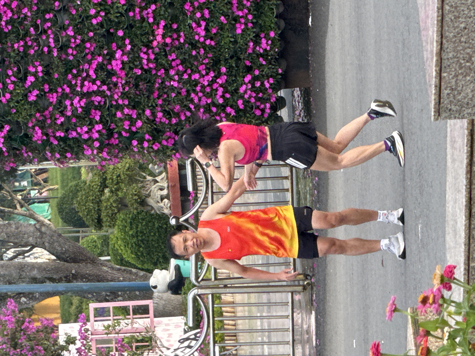 In the city of Lâm Viên - Đà Lạt, Vietnam, a man and a woman are captured in a dynamic outdoor scene. The man, wearing a vibrant orange and yellow sleeveless shirt with a flame-like pattern and black shorts, stands facing the camera with his left arm slightly extended. He has a neutral expression. Beside him, the woman, clad in a pink sleeveless crop top with black trim and black shorts, is captured mid-stride, her body angled away from the camera as she moves to her left. Her right arm is raised, touching her hair, and she appears to be looking to her left. Both individuals are wearing athletic shoes. They are positioned on an asphalt surface, with a metal fence and decorative potted plants in the background. A large wall covered in bright pink flowers dominates the right side of the frame, while trees and manicured bushes are visible further in the background. The scene appears to be set in a park or a recreational area, suggested by the presence of decorative elements and greenery.