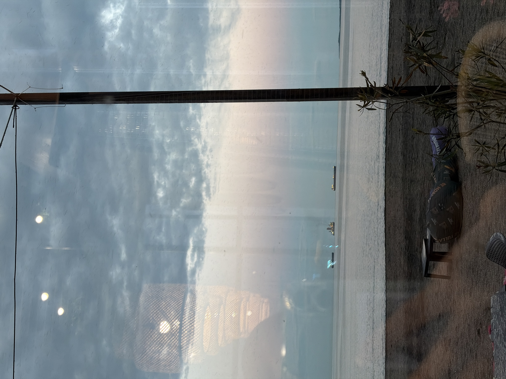 The image shows a view through a window, likely in Burgas, Bulgaria, overlooking the Black Sea on a cloudy day. The reflection in the window includes interior elements such as a hanging light fixture, a wooden beam and a potted plant. The foreground on the right side shows a portion of a patio area with a small black chair and possible part of a table. In the background, the sea is visible with what appear to be ships or distant buildings in the distance, with a cityscape visible on the horizon.