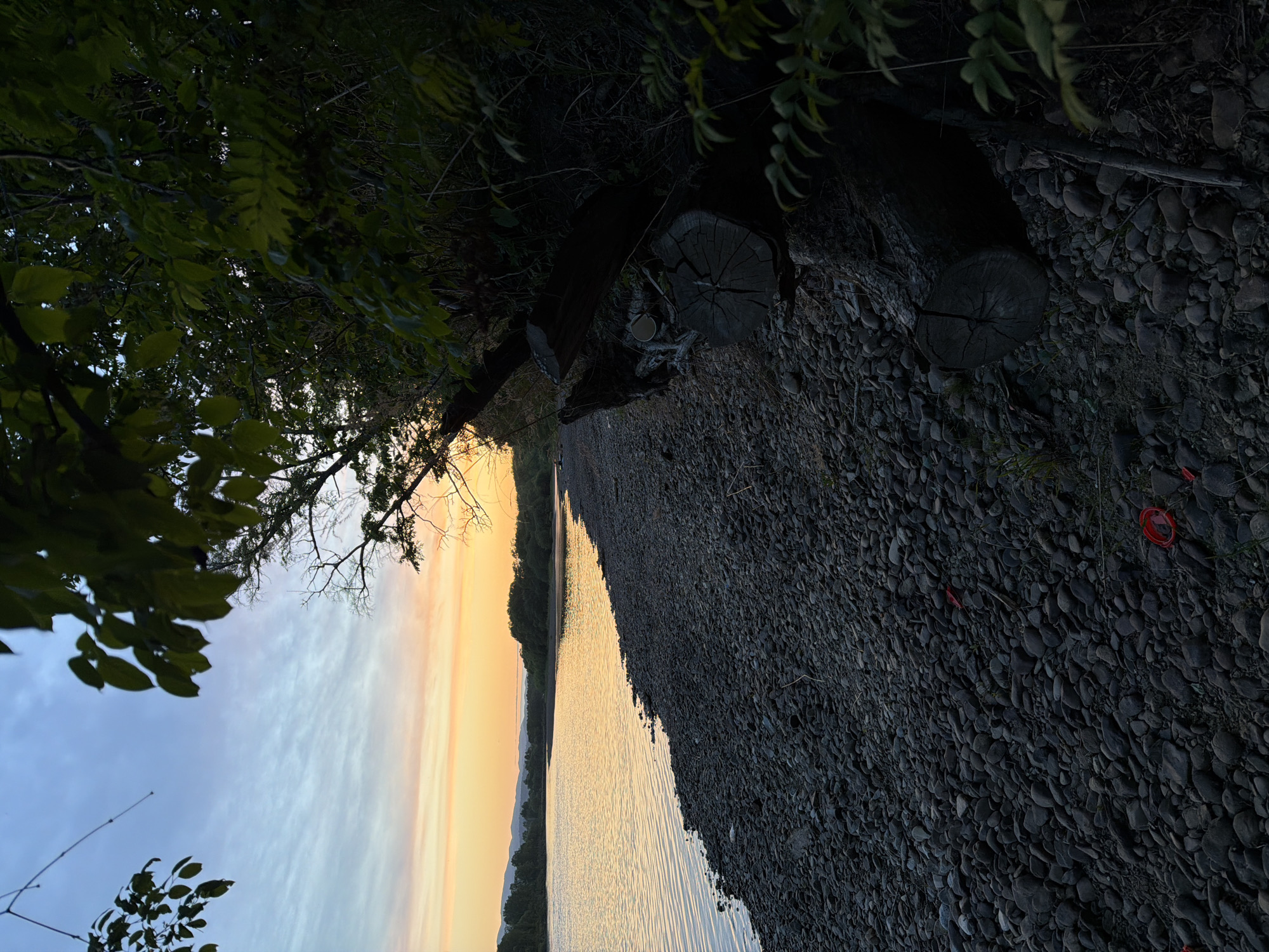 The image depicts a sunset over a riverbank. The left side of the frame shows the sky transitioning from blue to orange as the sun sets. Trees and foliage overhang from the top left corner, partially obscuring the sky. The river reflects the colors of the sunset, creating a golden hue on the water's surface. The right side of the frame is dominated by a rocky riverbank covered in dark, rounded stones. A cut tree trunk lies on the riverbank, along with scattered debris. The specific location is not discernible from the image. Birobidzhan, Russia, cannot be confirmed.