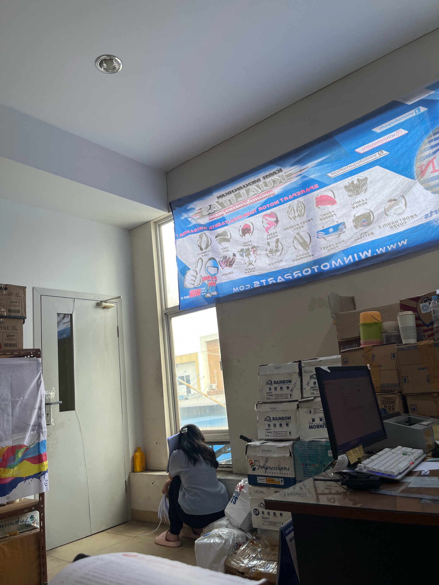 The image depicts an indoor scene, likely an office or storage area, with a woman sitting on the floor facing a window. She is wearing a light blue shirt and dark pants, with her hair pulled back. In front of her are stacks of boxes, some labeled with "RAINBOW" and "Impressions". A computer monitor, keyboard, and various office supplies are visible on a desk in the foreground. A large banner with blue and white text, partially obscured, is hung on the wall above the window. The room has fluorescent lighting and a white ceiling. The overall impression is one of a cluttered workspace. Without further information, it is impossible to definitively determine the exact location within Percut Sei Tuan, Indonesia, or the specific nature of the business.