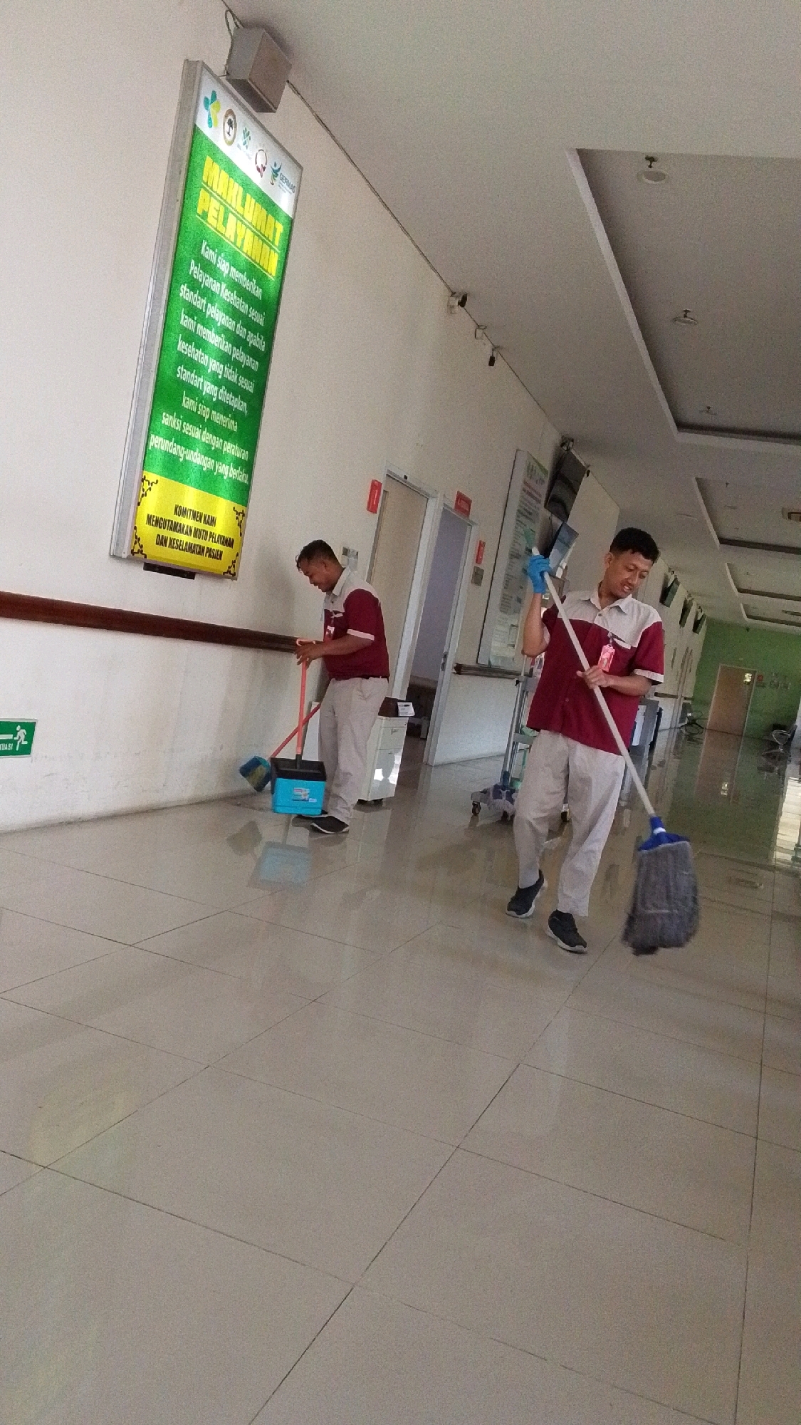 In the city of Pabelan, Indonesia, two male janitorial staff members are observed in a well-lit, tiled hallway of what appears to be a hospital or clinic. The hallway features light-colored walls and a dark brown handrail running along the left side. On the left wall, a large, illuminated green sign with text in Indonesian is prominently displayed.
The janitorial staff are dressed in matching uniforms consisting of a maroon polo shirt with beige accents and light-colored pants. The man on the left is sweeping the floor with a broom and dustpan. The man on the right is actively mopping the floor with a mop. Both individuals appear focused on their tasks.
The environment is clean and organized, with medical equipment such as an IV stand visible in the background. The overall impression is one of routine maintenance and cleanliness within a public facility.