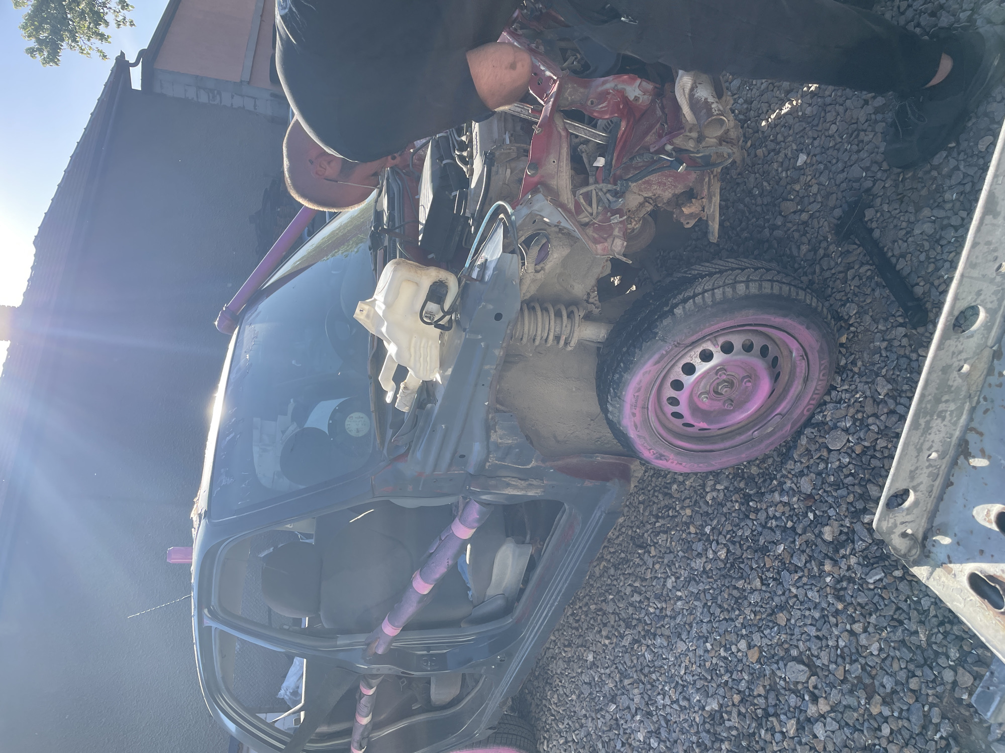 An outdoor scene in what appears to be a junkyard or vehicle repair area in Sofia, Bulgaria. The dominant subject is a heavily damaged car with the front end crushed and many components exposed. The car's body is primarily dark grey with pink accents on the roll cage and wheels. A man is working on the car, wearing dark clothing and shoes. The ground is covered in gravel. Other car parts are visible scattered around the scene, and the corner of a building is visible in the background.