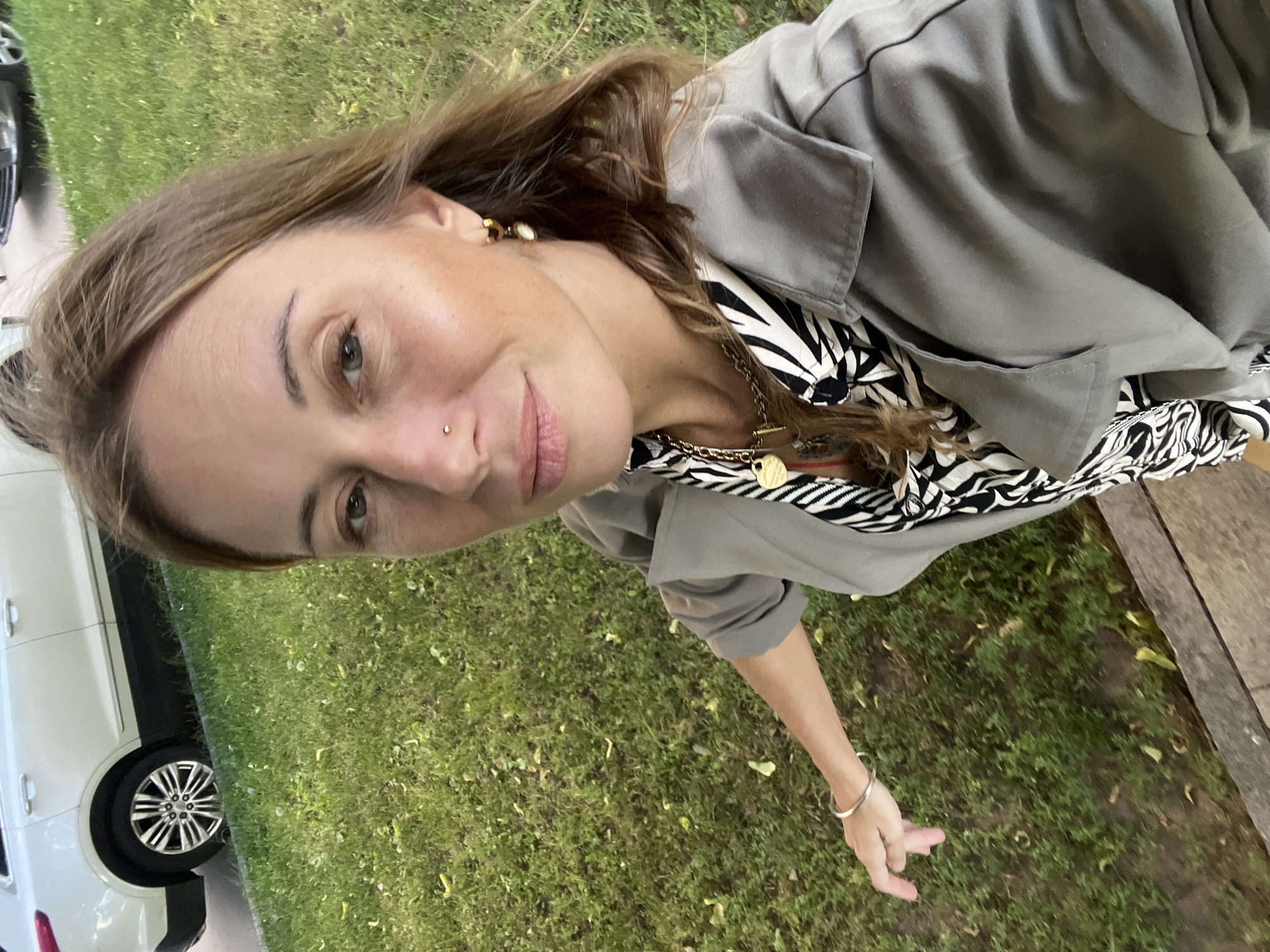 A woman with shoulder-length brown hair and a nose ring is captured in a selfie outdoors, with a blurred white SUV and green grass in the background. She is wearing a gray trench coat over a black and white patterned shirt and several gold necklaces. Her left arm is extended towards the camera, and she is wearing a silver bracelet. The location is a residential area with a paved sidewalk and grassy lawn.
