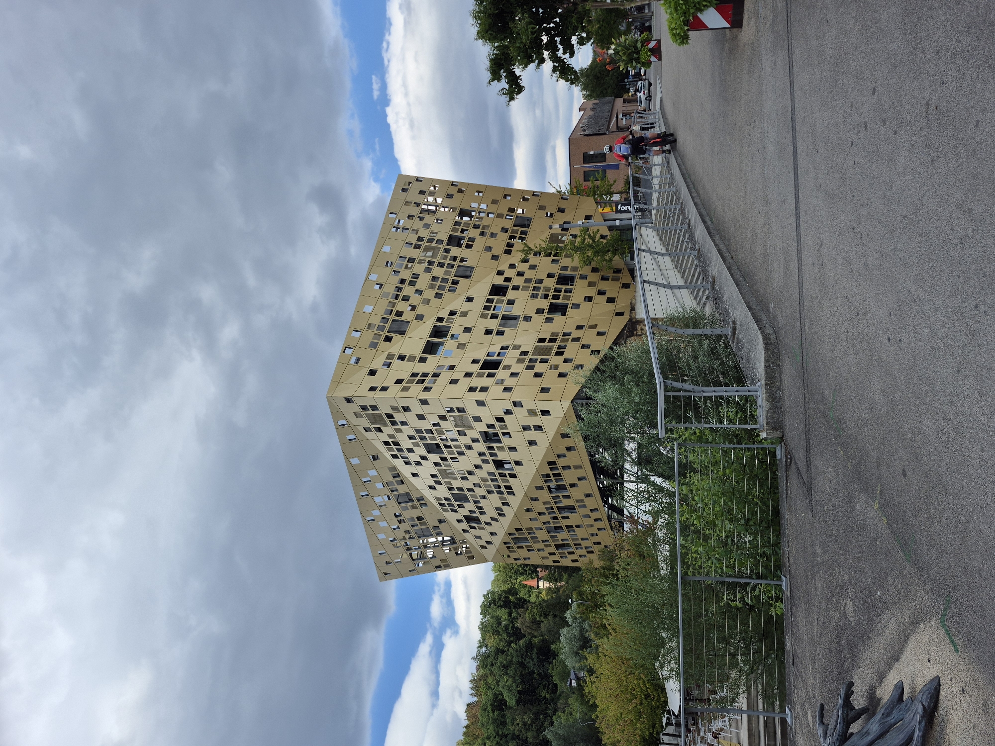A striking, gold-colored, angular building with a facade of irregularly placed square windows dominates the center of the frame. The building's geometric, almost cubist design is notable. In the foreground, a paved path curves to the right, bordered by a modern metal railing. Two individuals on bicycles are proceeding along this path, one in a red shirt and the other further ahead. Lush green trees and vegetation surround the area. In the background, a tree-covered hill rises, with a few other buildings visible, including one with a red turret. The sky above is a mix of grey clouds and patches of blue. The scene appears to be in a public outdoor space, possibly a park or a waterfront area within the city of Schwäbisch Gmünd, Germany.