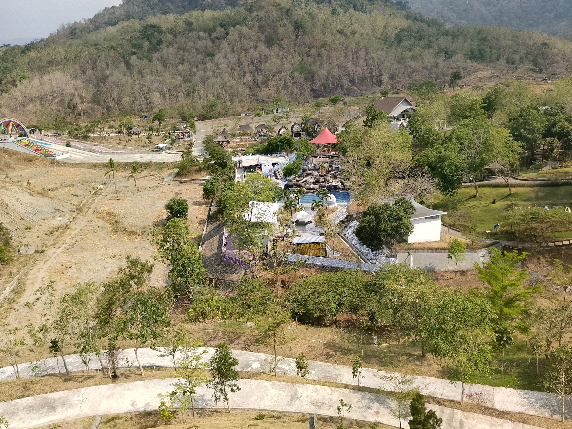 This is an aerial, high-angle, wide shot of a park with various recreational facilities situated in a hilly, arid environment. The park, located in Penanggapan, Indonesia, features a large rainbow-colored archway leading to a series of colorful water slides. Nearby, a complex of buildings and swimming pools is visible, including what appears to be a central pool with a rock waterfall feature. Several smaller structures, possibly huts or gazebos, are scattered throughout the park, some with red conical roofs. Green trees and vegetation are present, though much of the surrounding landscape is dry and brown, with sparse trees on the hills in the background. Paved pathways wind through the park, connecting different areas. The overall scene depicts a resort or amusement park designed to offer leisure and entertainment in a natural, hilly setting.