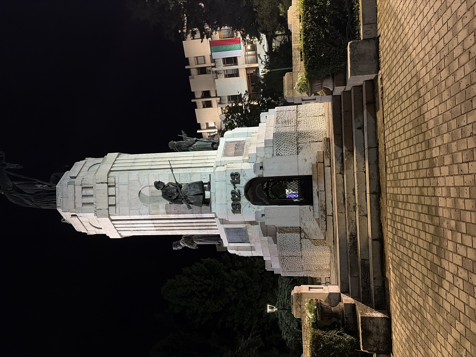 Captured at night in Veliko Tarnovo, Bulgaria, the image features the Monument to the Asenevtsi, a large, multi-tiered white stone structure adorned with bronze sculptures of historical figures. It stands at the edge of a park, with the Bulgarian flag displayed on a building in the background. A series of stone steps lead up to the monument, adjacent to a brick wall. The scene is illuminated by artificial lights, creating dramatic shadows and highlighting the details of the monument and surrounding environment.