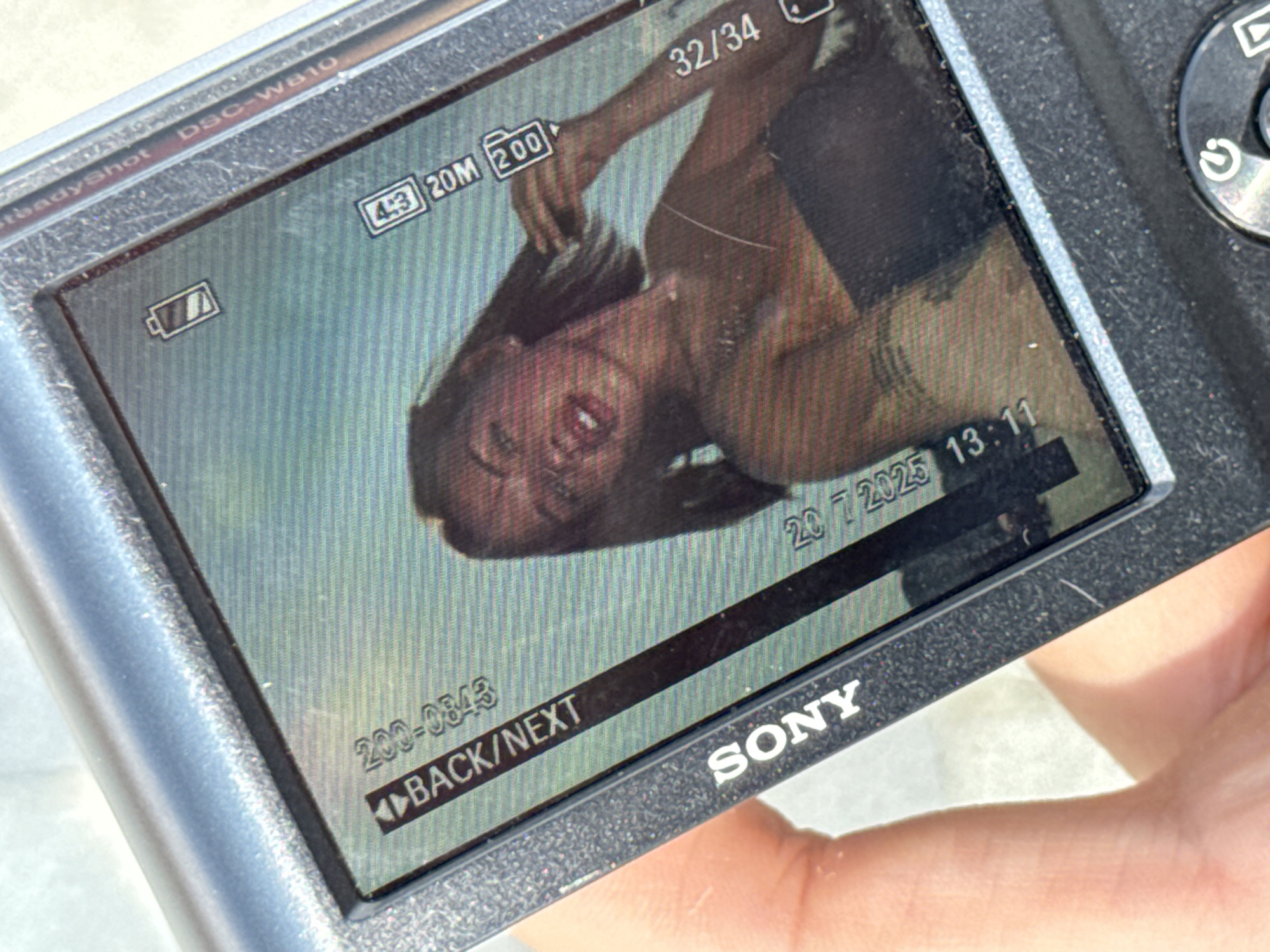 The image is a close-up photograph of a digital camera's LCD screen displaying a photo of a young woman. The woman has dark hair, which she is touching near her face. She is wearing a strapless top. The camera's display shows various information: a battery icon, "4:3 20M 200" indicating the aspect ratio, resolution, and zoom level, and "32/34" indicating the image number out of a total of 34. Additionally, the camera model "DSC-W810" and "SteadyShot" are visible along the top bezel. The bottom left of the screen shows the brand name "SONY" and the text "BACK/NEXT" with an arrow. The date and time "23.7.2025 13:11" are also displayed. The surrounding area visible in the photograph is a hand holding the camera, with a suggestion of an outdoor, bright environment. The image does not contain any information that would suggest a specific location in the city of Perm, Russia.