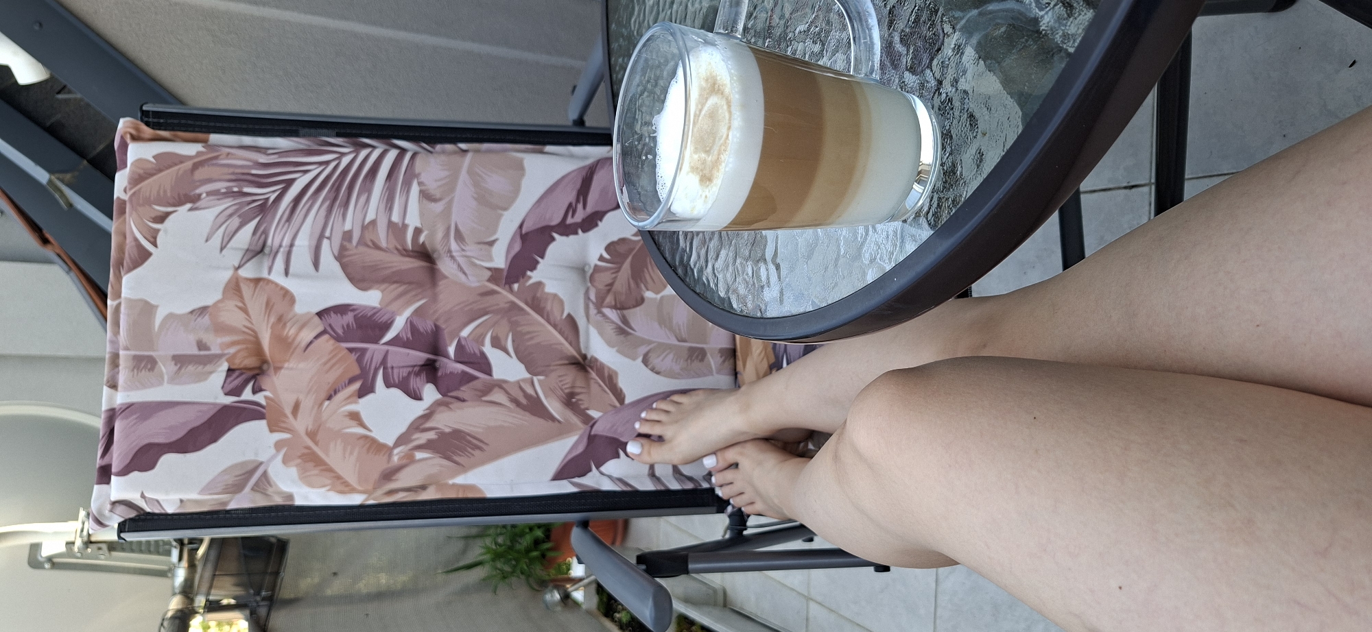 An eye-level, angled shot depicts a woman's legs and feet, a beverage on a glass-topped table, and a chair with a floral-patterned cushion, likely on a balcony. The woman's legs are fair-skinned and crossed at the ankles. Her toenails are painted white. A layered latte or similar beverage sits on the table. The chair cushion features a pattern of stylized brown and purple leaves. The background shows hints of an exterior setting with tiled flooring, plants, and a glimpse of another building. It is plausible that the location is Plovdiv, Bulgaria, but there are no distinguishing features that definitively confirm the city.