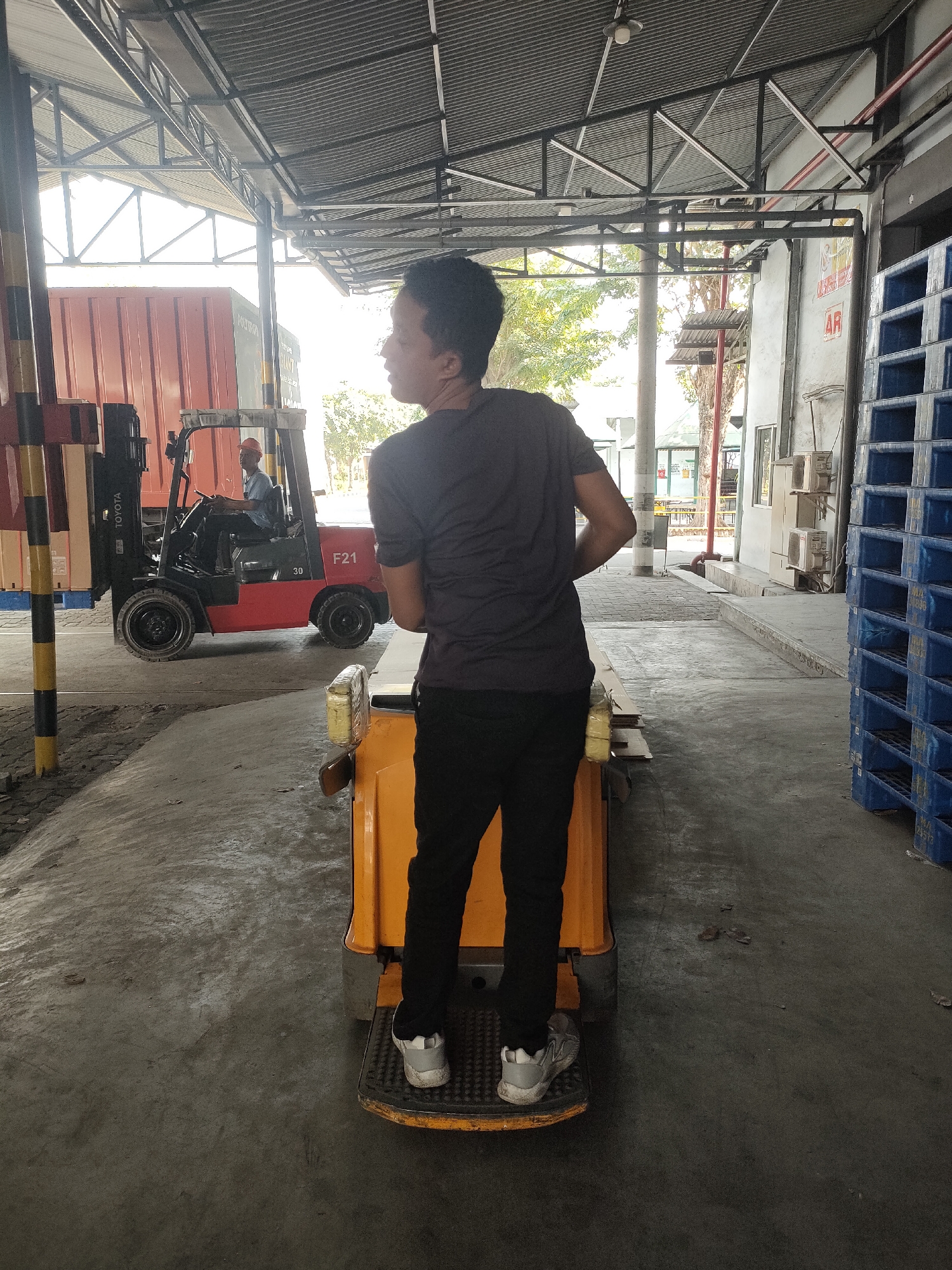 The image depicts a young man standing on an orange pallet jack in a warehouse setting in Sayung, Indonesia. He is wearing a dark t-shirt and black pants, with white athletic shoes. His back is to the camera as he appears to be looking at something to his left. In the background, a red Toyota forklift is being operated by another person wearing an orange hard hat and blue shirt. The forklift is positioned near a large orange shipping container. To the right of the image, stacks of blue plastic pallets are visible, and the environment suggests an industrial or logistics facility with a corrugated metal roof overhead and concrete flooring.