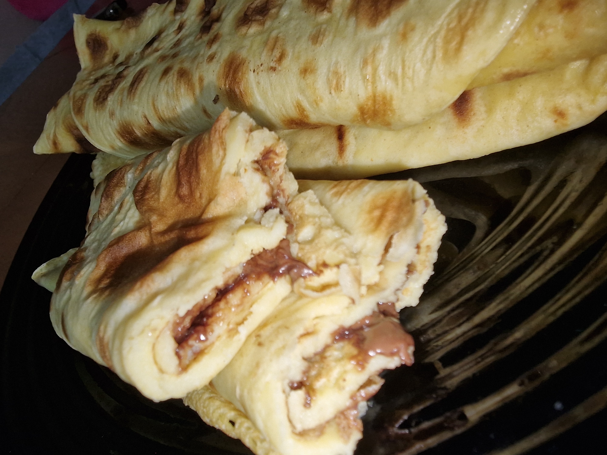 Here is a description of the image:
The image shows a stack of rolled crepes filled with a chocolate spread on a black plate. The crepes are golden brown with slightly darker browned spots, indicating they were cooked on a griddle or pan. The filling is a thick, dark brown spread, possibly Nutella or a similar chocolate-hazelnut spread. Some of the crepes are cut open, revealing the layers of crepe and filling. The plate is black and has a textured surface. The image is taken at a close range, focusing on the details of the crepes and the filling. The location is unknown.