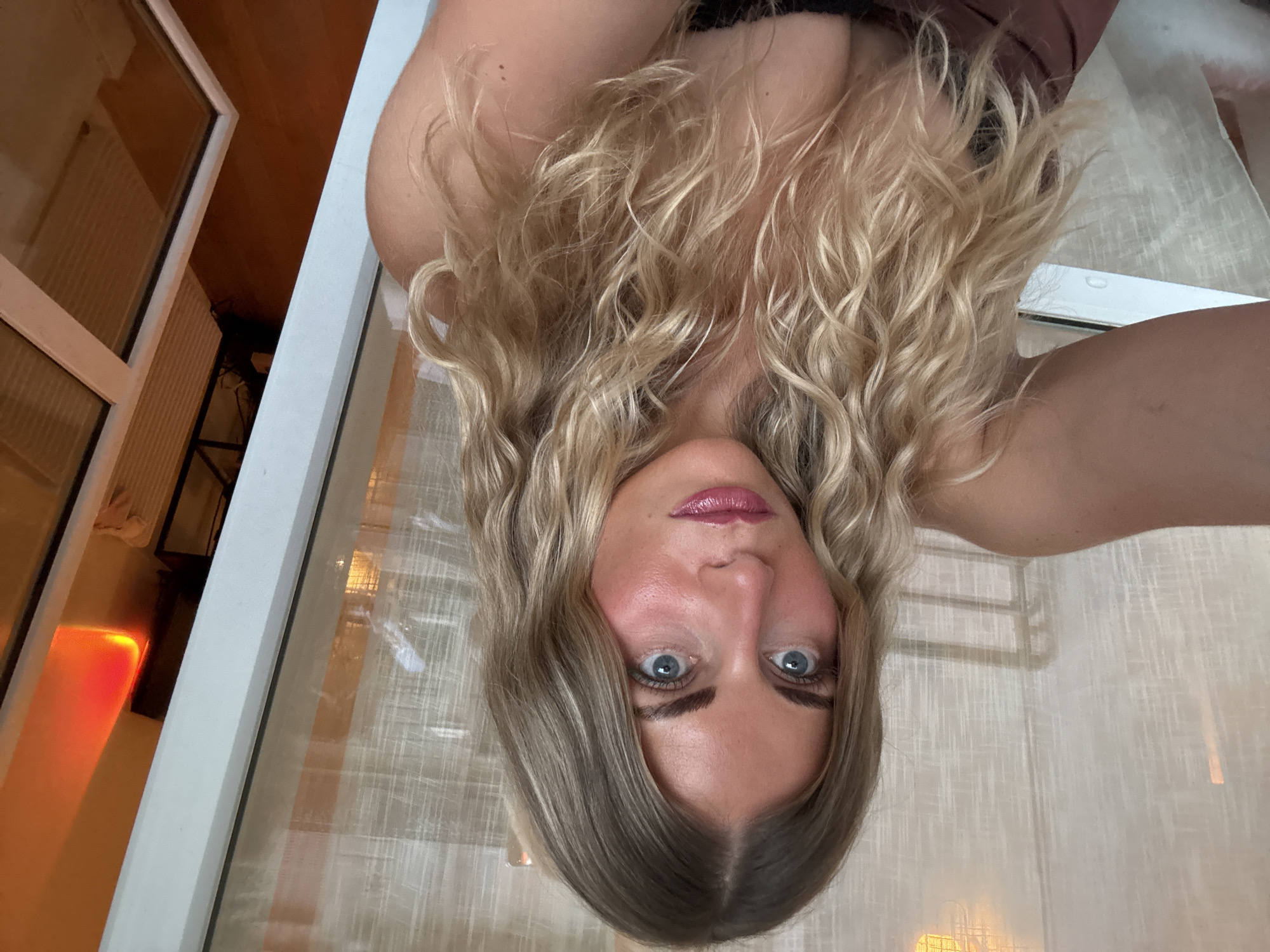A woman with blonde, wavy hair is positioned upside down in the frame, looking directly at the camera. Her eyes are blue. She has fair skin and is wearing what appears to be a brown top. Behind her is a window with white frames. The view through the window suggests an interior setting with warm lighting, possibly a hotel room. It is impossible to determine if the location is Paris, France from the given image.