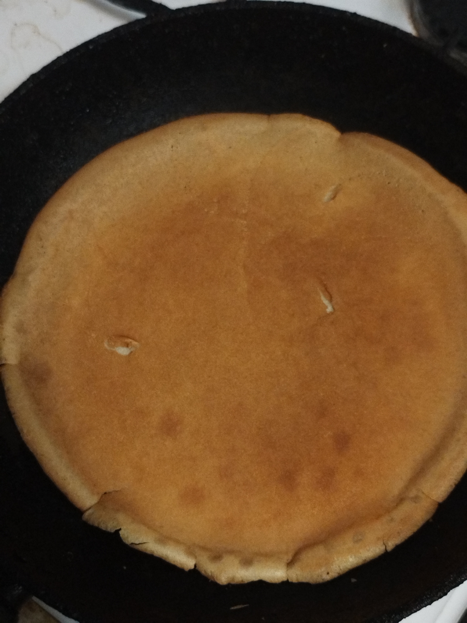 A close-up, overhead shot shows a large, golden-brown pancake cooking in a black skillet. The pancake fills most of the frame, with its slightly uneven edges resting against the sides of the pan. The surface of the pancake is mostly smooth, with a few small, lighter-colored bubbles or imperfections visible. The lighting appears to be indoor, with a slight glare on the pancake's surface. The background is out of focus, showing a white, speckled surface and hints of the skillet's handle. The scene is simple and domestic, suggesting a breakfast or brunch preparation. There is no visible text.