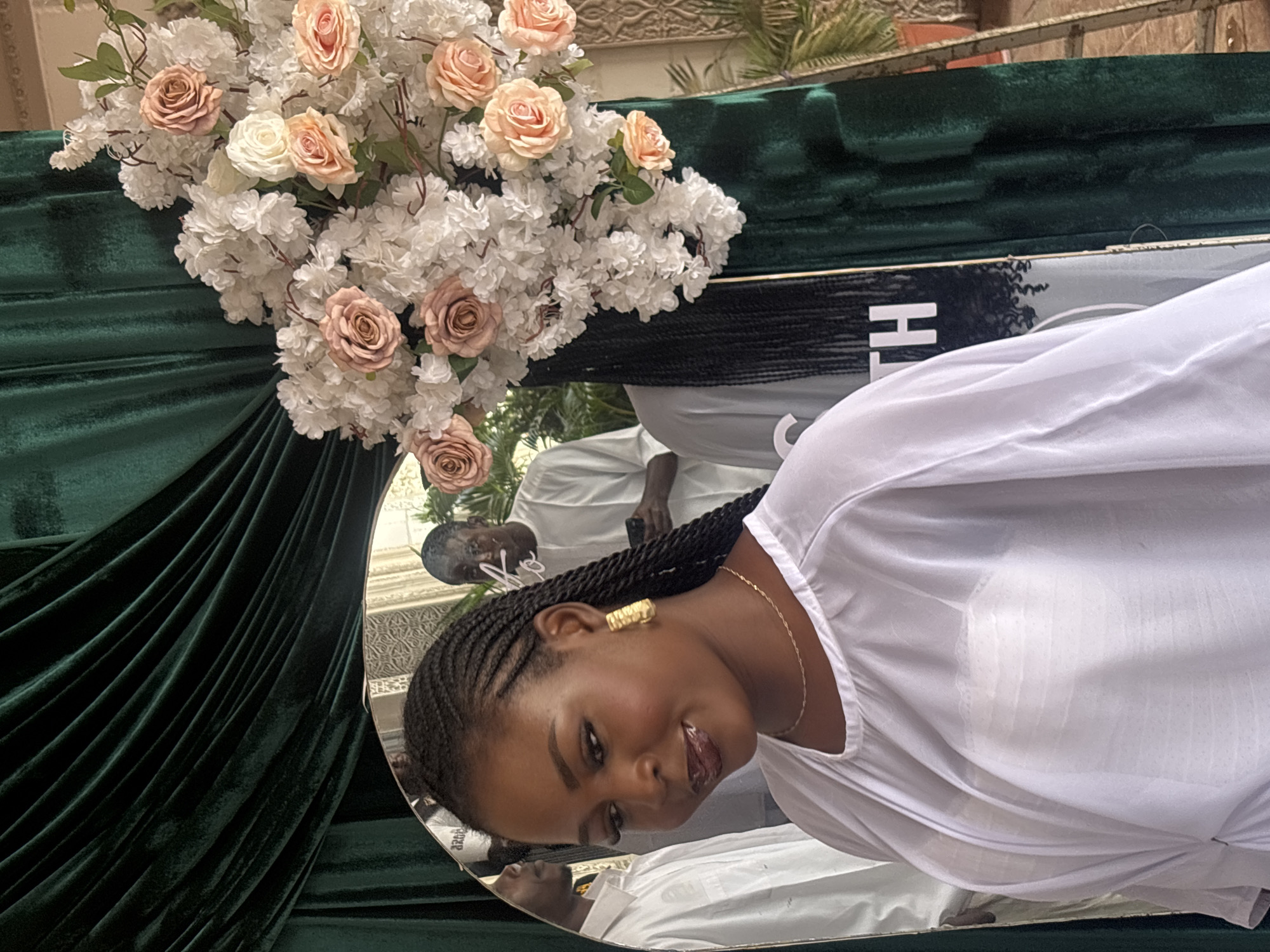 The image shows a woman in a white dress, with braids and gold earrings, looking into a mirror. She is standing in front of a floral arrangement and a green velvet backdrop, possibly at an event. In the reflection, other people in white attire are visible. The setting appears to be indoors or a covered outdoor space, given the lighting and backdrop. There is text visible on a banner behind the floral arrangement, though only the letters "TH" are clearly discernible. The overall atmosphere suggests a formal occasion or celebration. The time of day is not explicitly indicated but the lighting appears to be natural, suggesting daytime. The location is specified as Jos, Nigeria.