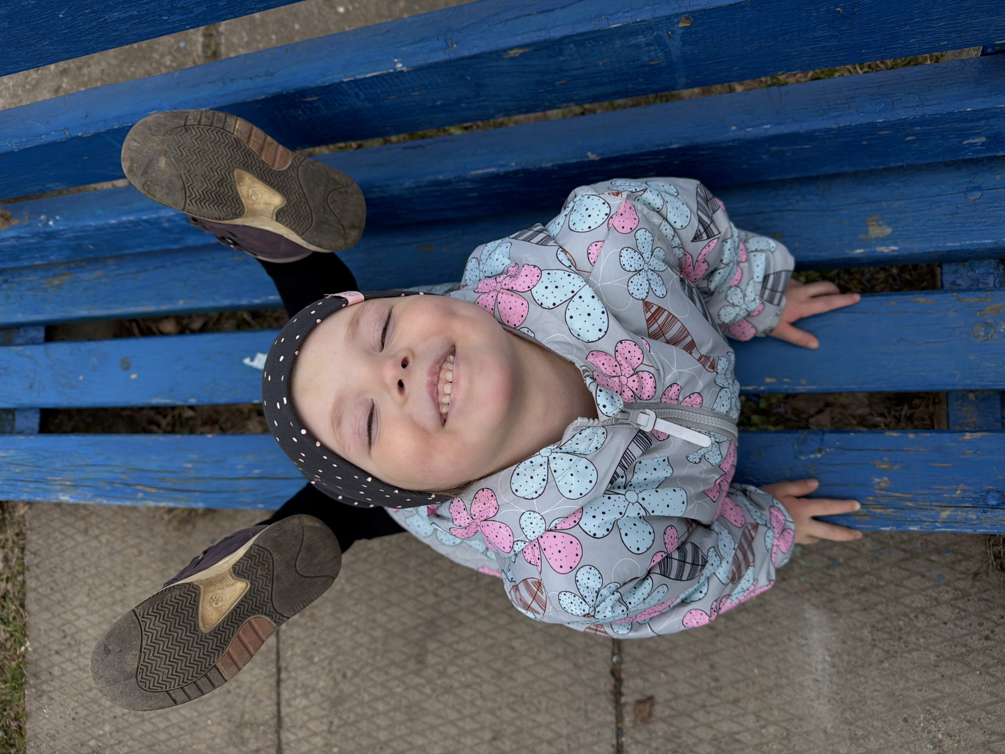 A young child lies on a blue, wooden bench outdoors, likely in a park or public space in the city of Votkinsk, Russia. The child wears a light gray jacket with a floral pattern of pink and light blue flowers, a black polka-dot headscarf, and black leggings. The child lies face-up on the bench with the legs extended upward, feet in the air. The bottoms of the child's dirty, dark-colored shoes are visible. The bench is made of multiple horizontal wooden planks painted blue. The area surrounding the bench appears to be paved. The child's hands are flat against the bench. The child smiles, showing teeth.