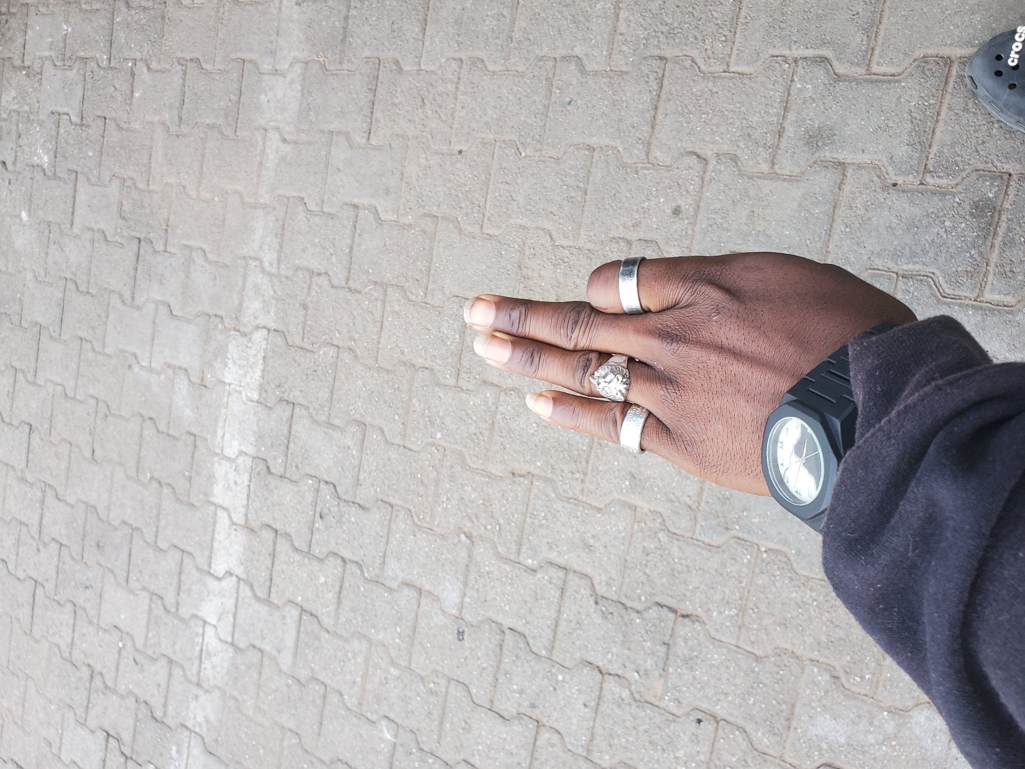 The image shows a close-up view of a person's hand and wrist against a backdrop of interlocking grey paving stones, typical of urban outdoor spaces in cities like Jos, Nigeria. The hand is adorned with two silver rings, one on the index finger featuring a lion's head design and a plain band on the middle finger. The wrist sports a black wristwatch with a round, white analog dial and a textured black strap. The person is wearing a dark blue or black long-sleeved garment, likely a jacket or sweatshirt. The fingers are extended upwards, with the fingernails visible. The paving stones are laid in a regular pattern, and the lighting suggests an outdoor environment, possibly a sidewalk or plaza.