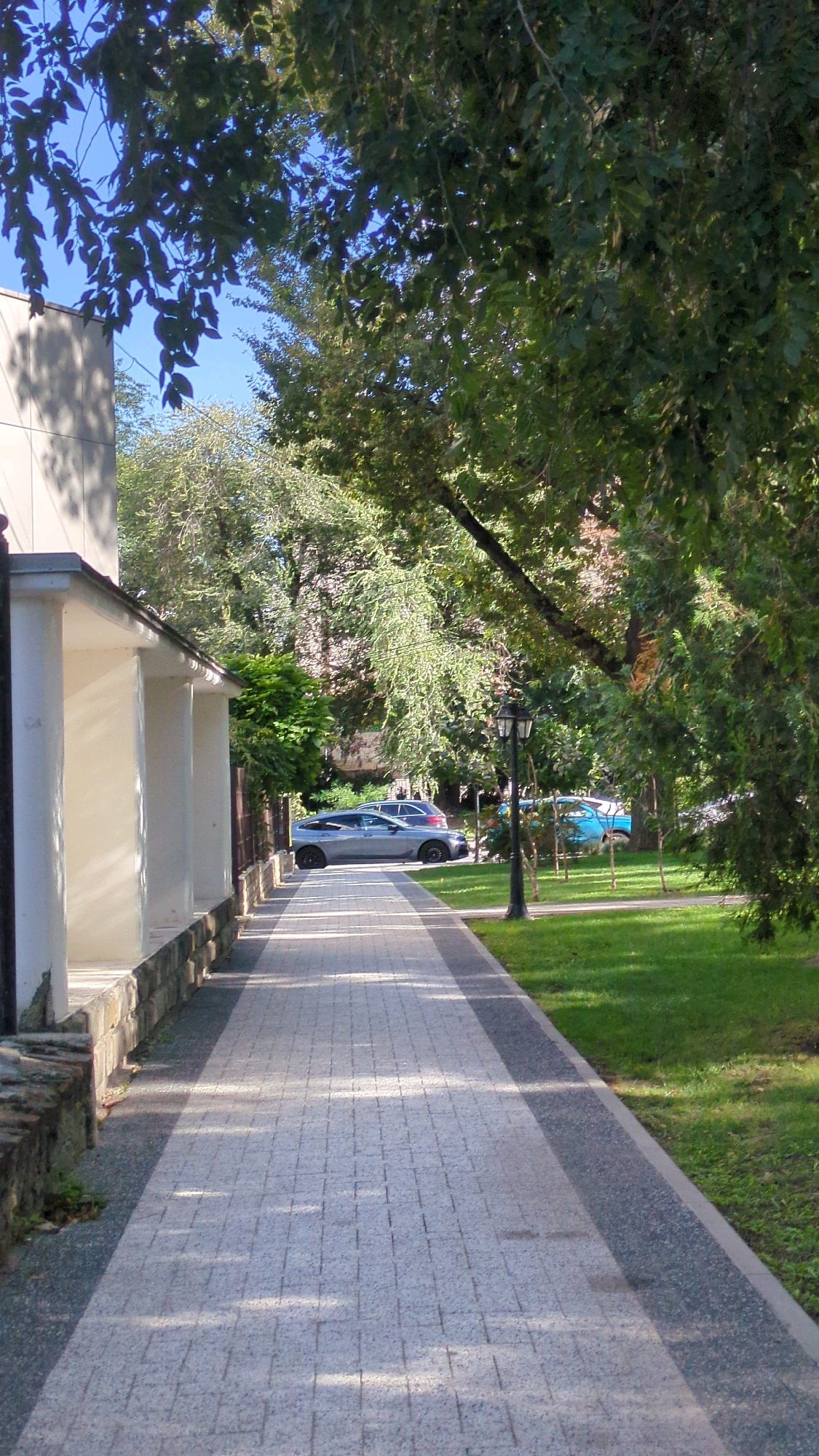 The image shows a paved walkway in a park-like setting in Chișinău, Moldova. The walkway is made of light-colored cobblestones, bordered by darker paving stones and a strip of green grass. To the left of the walkway is a white building with a colonnade and a low stone wall. Sunlight casts shadows across the walkway and onto the building. Several cars are parked along the side of the walkway, and a street lamp stands near the center of the path. Lush green trees with dense foliage line the right side of the walkway and the background, obscuring the sky in places. The overall impression is a peaceful, well-maintained urban park.