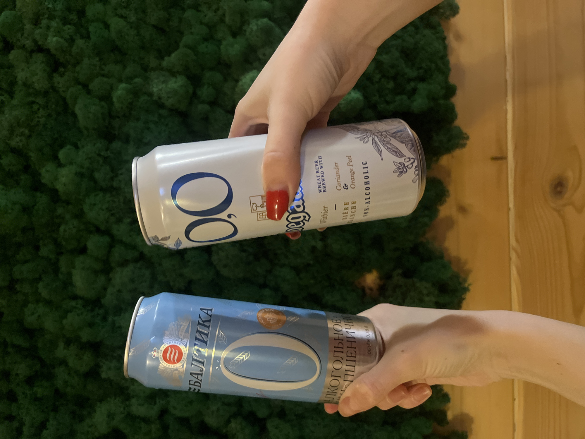 The image shows two hands holding canned non-alcoholic beers in front of a wall covered in artificial green moss. The upper hand is holding a white can labeled "Hoegaarden Witbier 0.0%" while the lower hand holds a light blue can labeled "Baltika Non-Alcoholic Wheat." The cans are positioned parallel to each other and appear to be presented for display. A portion of a wooden panel is also visible on the right side.
