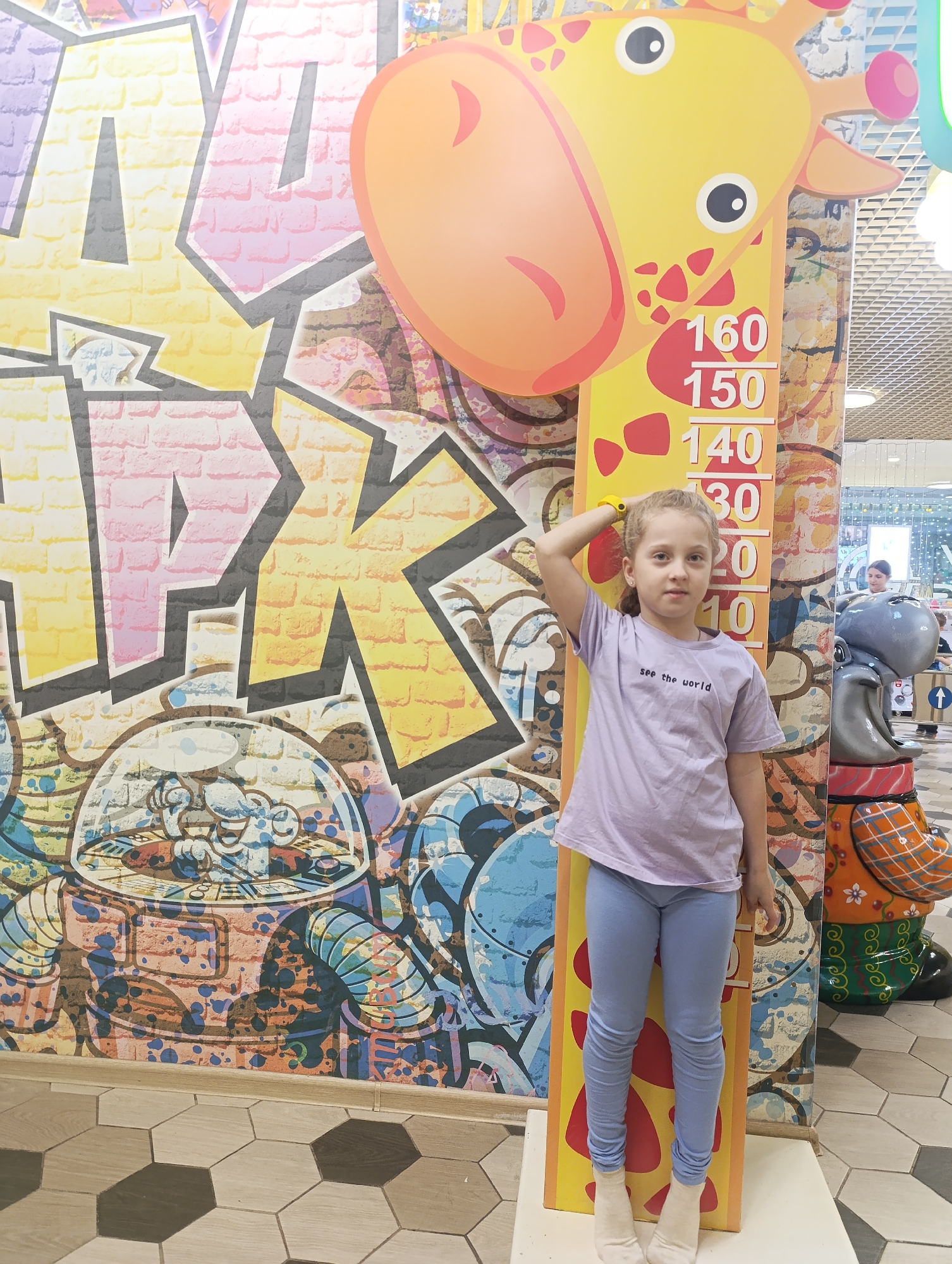 A young girl stands next to a giraffe height chart in an indoor location, possibly a shopping mall or children's play area. The girl has light blonde hair and is wearing a light purple t-shirt with the words "see the world" and light blue leggings with white socks. She is touching her head with her right hand, measuring her height against the giraffe chart. The chart is a yellow and orange giraffe with height measurements marked in centimeters. Behind the girl and the chart is a colorful graffiti-style mural with cartoon-like images and brick patterns. To the right of the girl is a statue of a hippopotamus with a painted design. The lighting is bright and the environment appears to be a public space with other individuals visible in the background. The image was taken in the city of Unknown, Unknown.