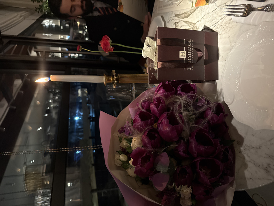 The image depicts a dimly lit, upscale dining table scene at night, identified as likely being within the Hermitage Restaurant in Yerevan, Armenia, based on the branding visible. A large bouquet of deep purple peonies and white roses, wrapped in light purple paper, occupies the left side of a marble-patterned tabletop. Centrally, a lit white taper candle stands in an ornate gold-colored candlestick, positioned next to a clear drinking glass. To the right of the candle, a small, open, dark brown gift bag from "HERMITAGE PERFUMES & COSMETICS" is visible, containing white tissue paper. Adjacent to the gift bag, two red ranunculus flowers are presented in a slender vase. A white ceramic plate with a decorative rim is placed in the right foreground, accompanied by a fork resting on a white napkin. Partially visible on the far right, a man with a beard, wearing a dark jacket and a striped tie, is seen looking down at a menu or document. The background features large windows overlooking blurred city lights at night.