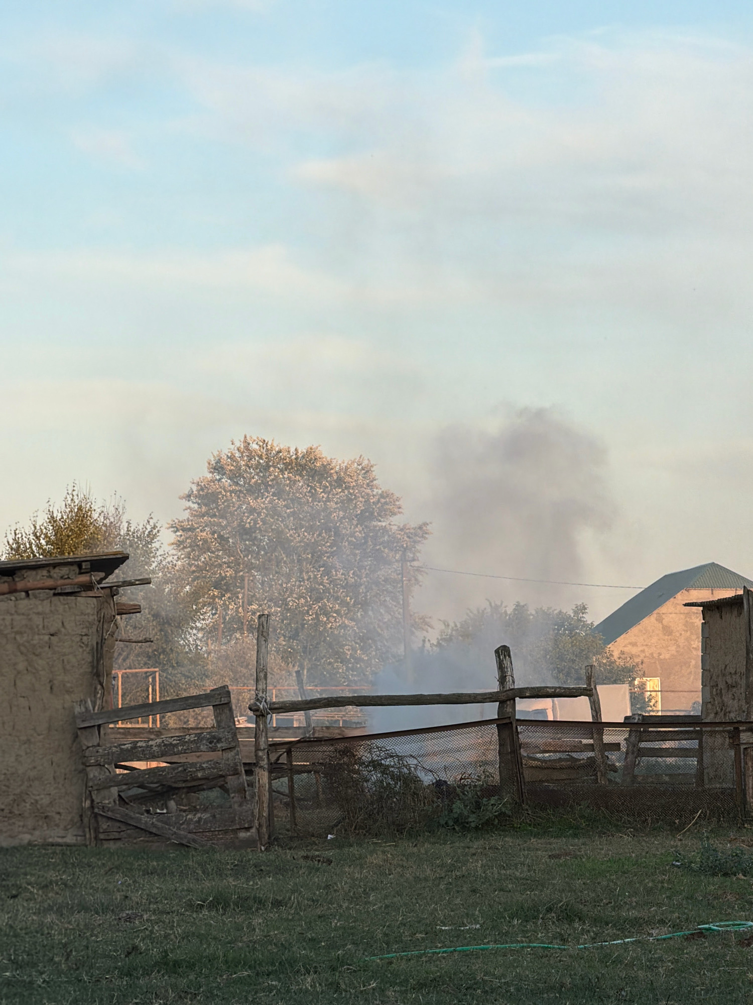A scene captures a rural setting with a dwelling, fences, and smoke rising into the sky. The sky is a pale blue with faint clouds. In the foreground, green grass covers the ground, with a green hose visible near the bottom. A rustic wooden fence encloses a yard area. Beyond the fence, there is a large tree with light-colored foliage and other trees. To the right, a building with a green roof is visible, partially obscured by smoke. A smaller structure, possibly made of adobe, is on the left. Smoke billows from behind the fence and trees, obscuring parts of the landscape and suggesting a fire or burning activity. There are no people or animals visible in the image. The scene appears to be from an outdoor location in a rural area, possibly a farm or village.
