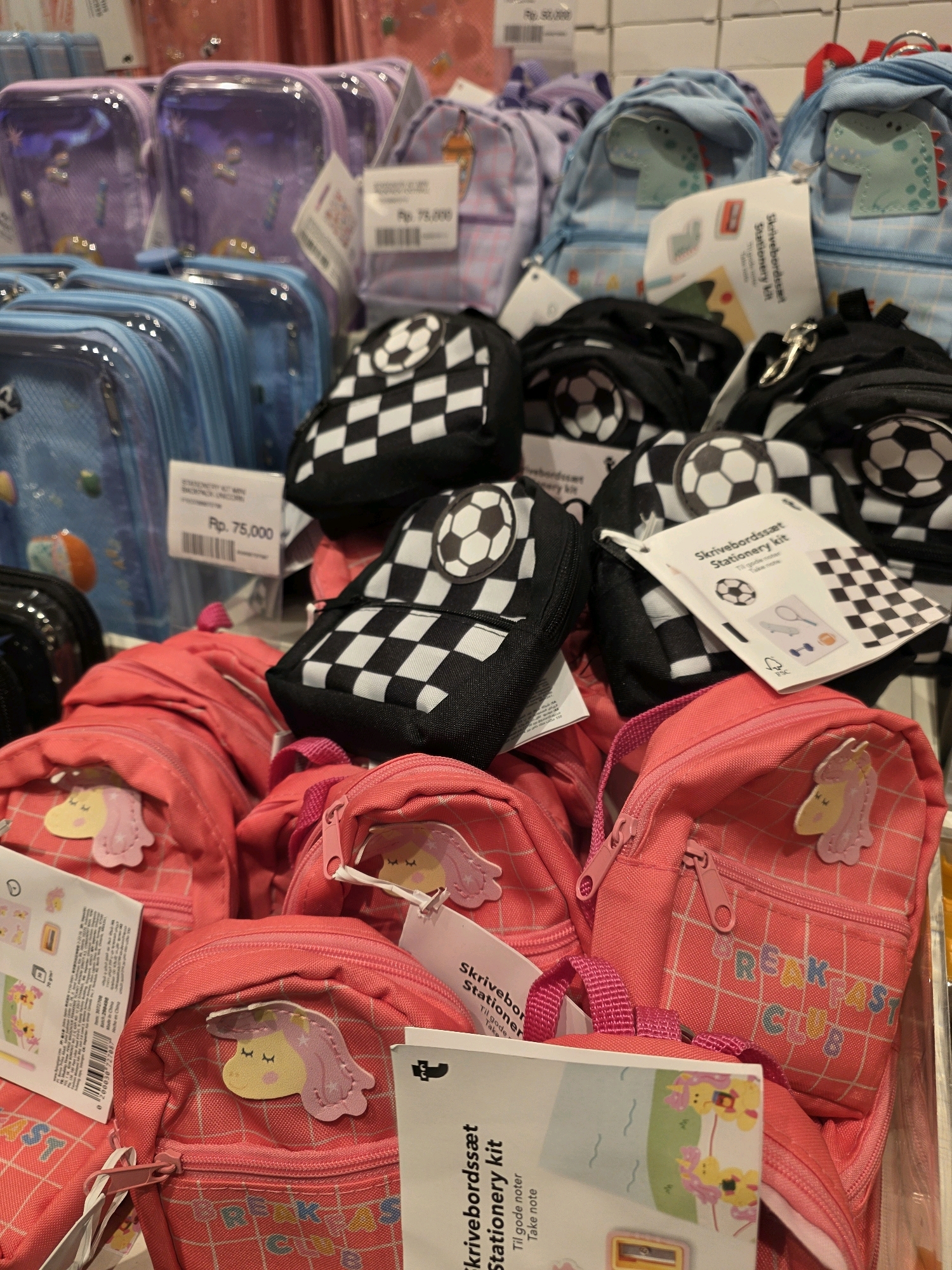 This is a close-up, eye-level shot of a retail display of various small backpacks and stationery kits. The products are primarily for children and feature bright colors and playful designs. In the foreground, several small pink backpacks with unicorn appliques are prominently displayed. Behind these, a selection of black backpacks adorned with a checkerboard pattern and soccer ball designs are visible. Further back, lighter blue backpacks with dinosaur appliques and translucent purple pouches are also part of the display. Some of the items have price tags and product labels attached. The overall impression is of a neatly arranged collection of merchandise. The scene is likely a store in Bandung City, Indonesia, given the visible price notations in "Rp.", which is the Indonesian Rupiah.