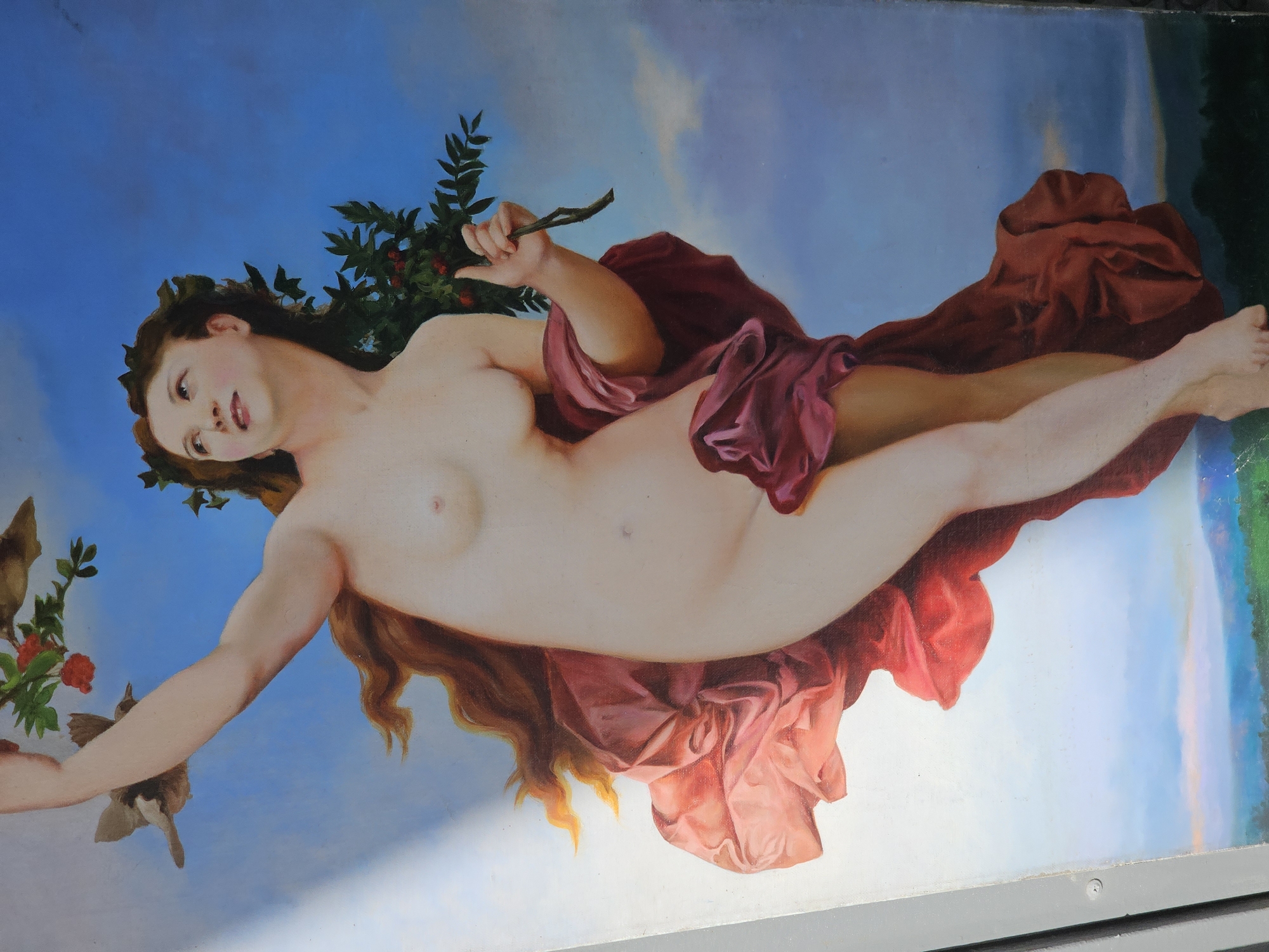 The image depicts a classical-style painting of a nude woman reclining against a blue sky backdrop. She has fair skin, reddish-brown hair adorned with leaves, and is partially draped with red fabric. In one hand, she holds a branch with berries; in the other, she holds a branch with leaves that has two birds perched on it. The painting is located in the city of Moscow, Russia, though the specific location within the city is not readily apparent. The painting appears to be mounted on a wall or structure, with a gray frame visible along the lower edge.