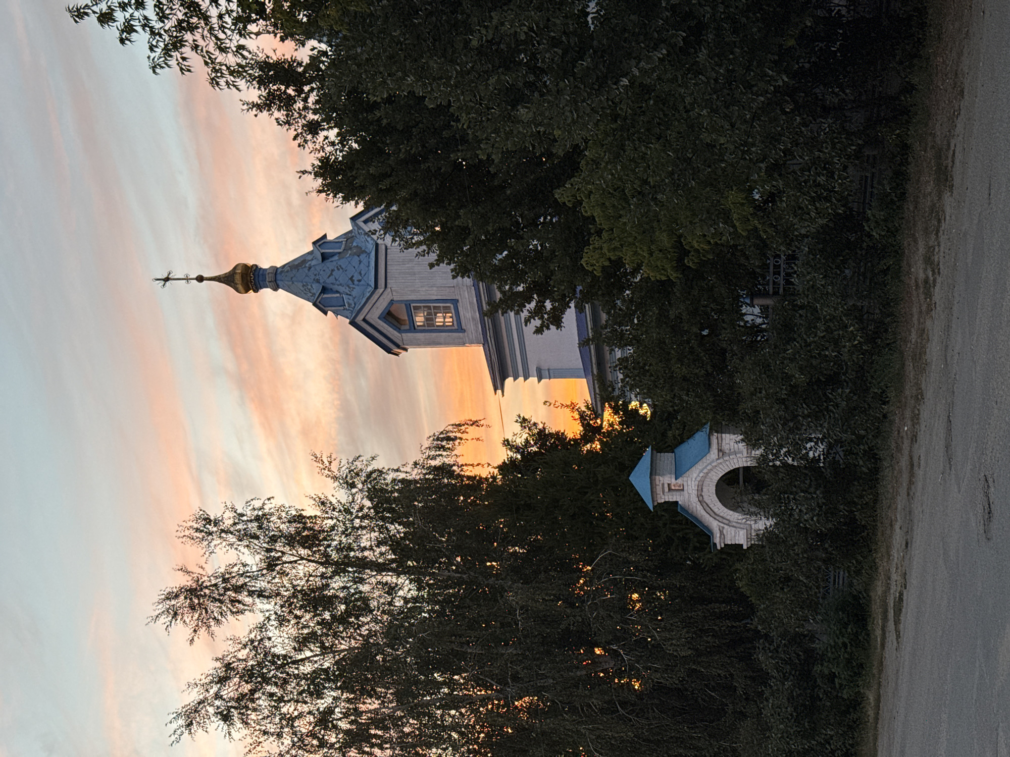 The image depicts an Orthodox church in the city of Sol-Iletsk, Russia, during sunset. The sky is a mix of soft pinks, oranges, and blues, with wispy clouds. A prominent feature is the church's blue-domed bell tower, topped with a golden cross, which is partially obscured by dark green foliage. To the left, a large, leafy tree with dark branches dominates the frame. In the foreground, a small, arched gateway or chapel with a blue roof and white brick construction stands amidst dense green bushes. A paved road or path runs horizontally across the bottom of the image, with a few patches of dry grass and some unevenness on its surface. The overall scene is one of tranquility and religious architecture set against a colorful twilight sky.