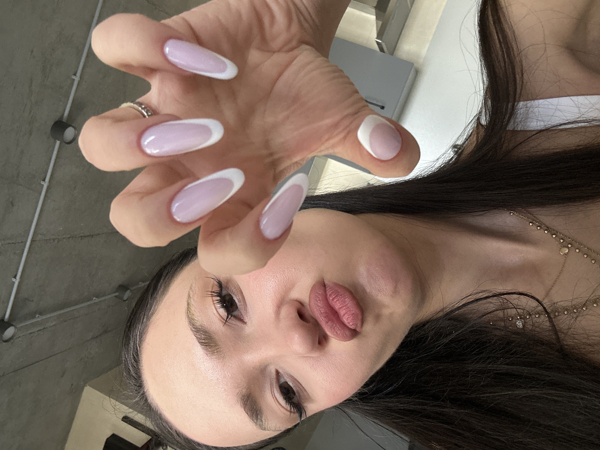 A woman is holding her hand towards the camera, showing off her long, oval-shaped nails with a French manicure style – a pale pink base and a thin white tip. She is looking up at the camera with a slight pout. She has dark hair. She is wearing a white top and a gold necklace. The background shows a gray concrete ceiling with exposed pipes and a glimpse of a gray desk or cabinet. It is not possible to confirm the location in Zovuni, Armenia based on the image.