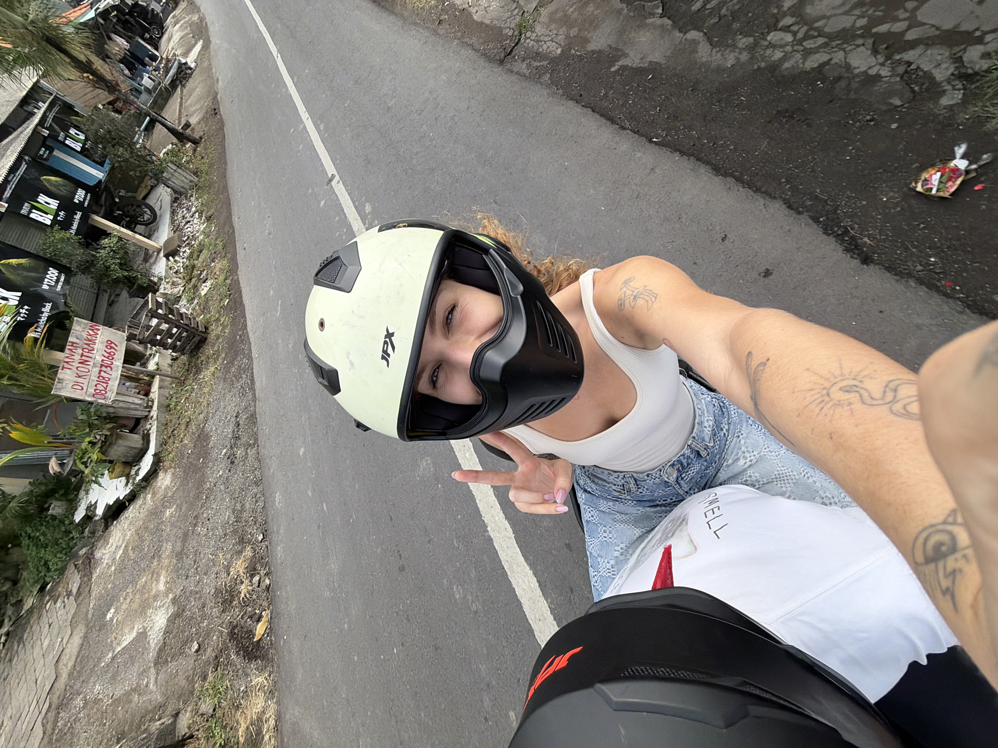 A woman wearing a white tank top, blue patterned jeans, and a light-colored helmet with a black face shield is riding on the back of a motorcycle in Mengwi, Indonesia. She is smiling and holding up two fingers in a peace sign. Tattoos are visible on her arm. The road is asphalt with a white line marking the lane. A second, black helmet is visible in the foreground, to the left. In the background, there is a building with signage that appears to be in Japanese and English, and a palm tree. There is also a sign in Indonesian that reads "TANAH DI KONTRAKKAN" which translates to "LAND FOR RENT." The environment appears to be a roadside setting.