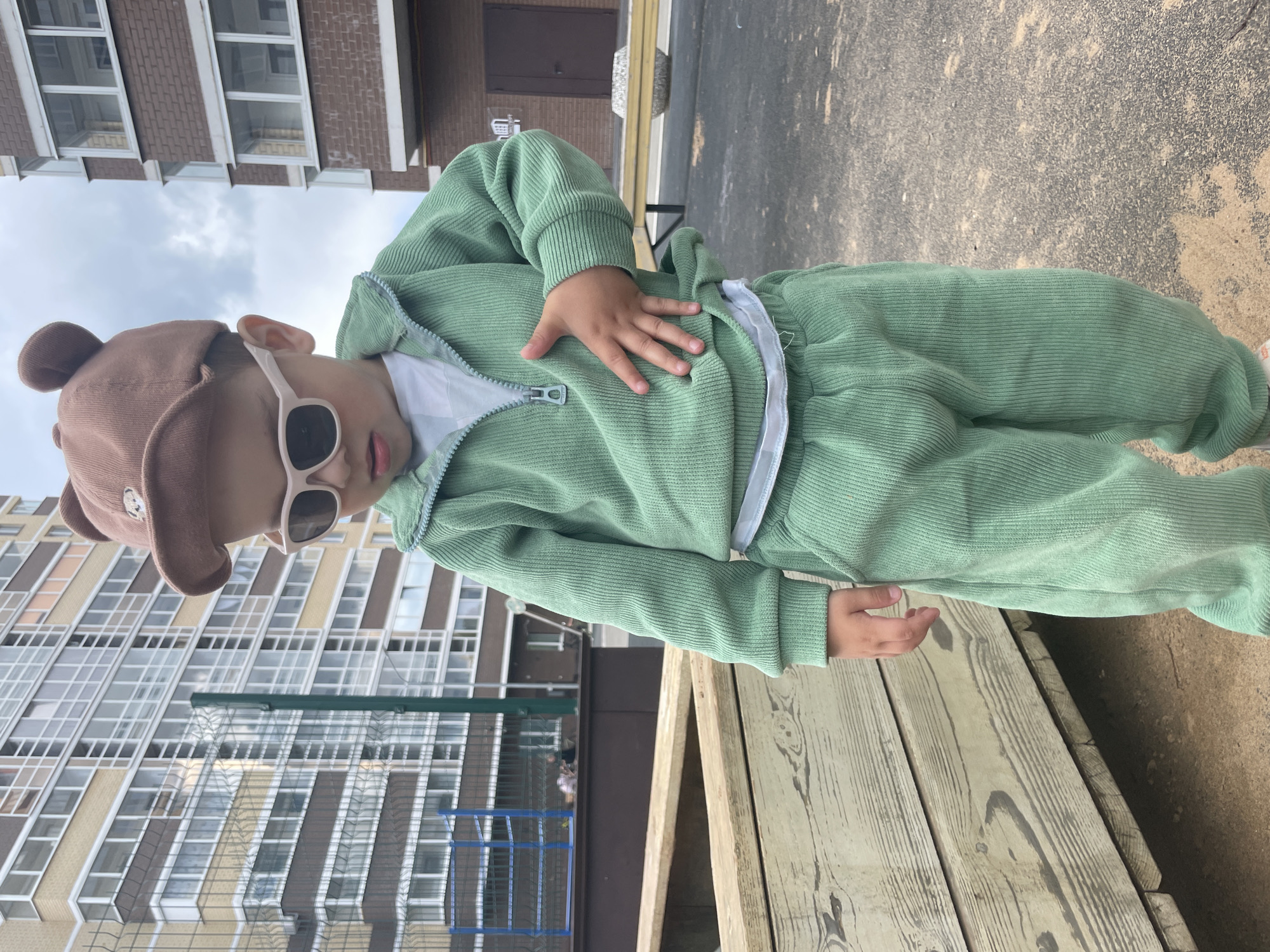 A child, wearing a light green corduroy tracksuit, a brown hat with small bear ears, and white-framed sunglasses, stands on weathered wooden planks outdoors. The child's left hand rests on their chest. Behind the child is a multi-story building with many windows, characteristic of urban architecture. The ground visible includes sections of asphalt and sandy dirt. It is daytime, and the lighting suggests partially cloudy weather. The location is in Izhevsk, Russia.