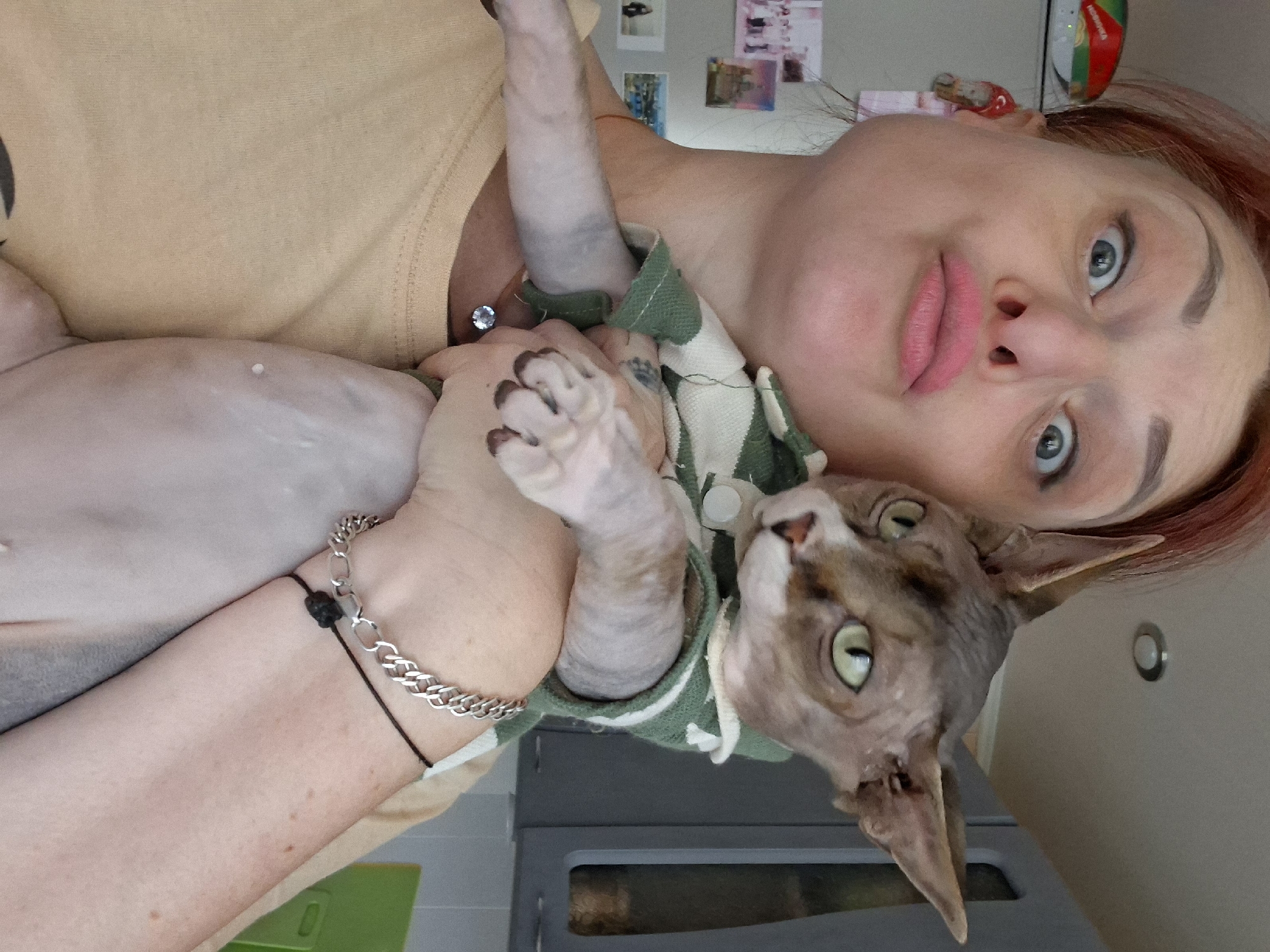 A woman with reddish-brown hair and wide, surprised eyes is holding a hairless cat. She is wearing a light yellow t-shirt. The cat is wearing a green and white striped sweater. The woman has her lips pursed in a pout. She is wearing a silver chain bracelet and a thin black bracelet with a knot. A clear gem earring is visible on her left earlobe. The background appears to be a kitchen with grey cabinets. The image is a close-up, selfie-style photograph. The location of Myadzyel, Belarus is not discernible from the visual information provided in the image.