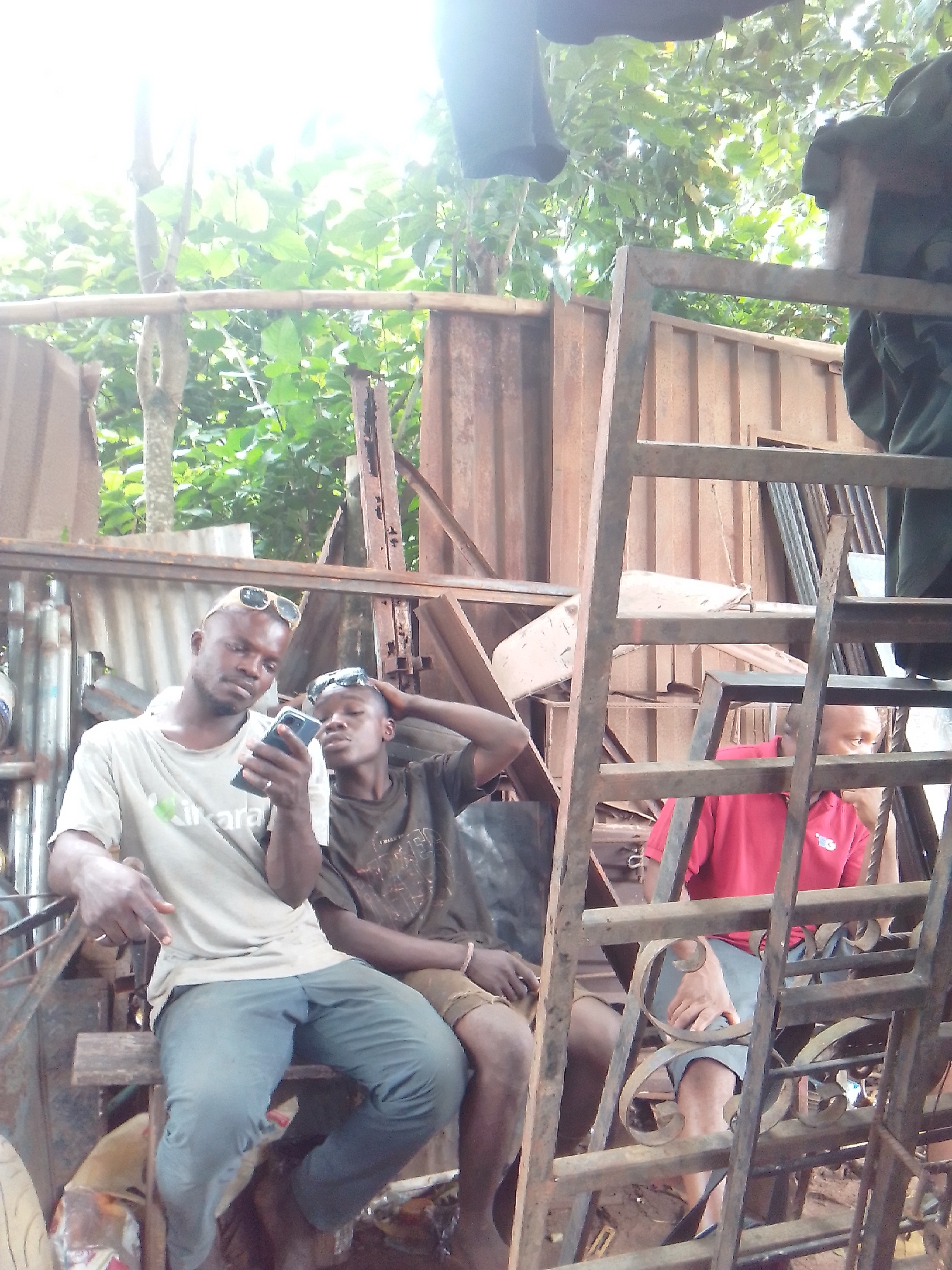 The image depicts three men gathered in an outdoor, somewhat cluttered environment under bright daylight. In the foreground, to the left, a man with a beard and sunglasses pushed onto his head wears a light-colored t-shirt with green text, "Ikara" or "Hikara," visible across the chest. He is holding a smartphone and intently looking at its screen, with his right hand resting on his leg.
Seated closely beside him, another man with sunglasses on his head wears a dark t-shirt and shorts. He also appears focused on the smartphone held by the first man, with one hand resting on his head. Both are sitting on a makeshift wooden or metal structure.
To the right and slightly behind these two, a third man is partially visible, wearing a red polo shirt with a small, indistinct logo on its right chest. He is seated behind a framework of rusty metal bars, possibly part of a ladder or scaffolding, which dominates the right side of the frame.
The background features a mix of corrugated metal sheets, various metal scraps, and lush green foliage from trees, suggesting an informal workshop, scrapyard, or a casual gathering spot in a semi-urban or rural setting. The bright, almost overexposed lighting, particularly from the top left, indicates a sunny day. The men appear relaxed and engaged with the smartphone.