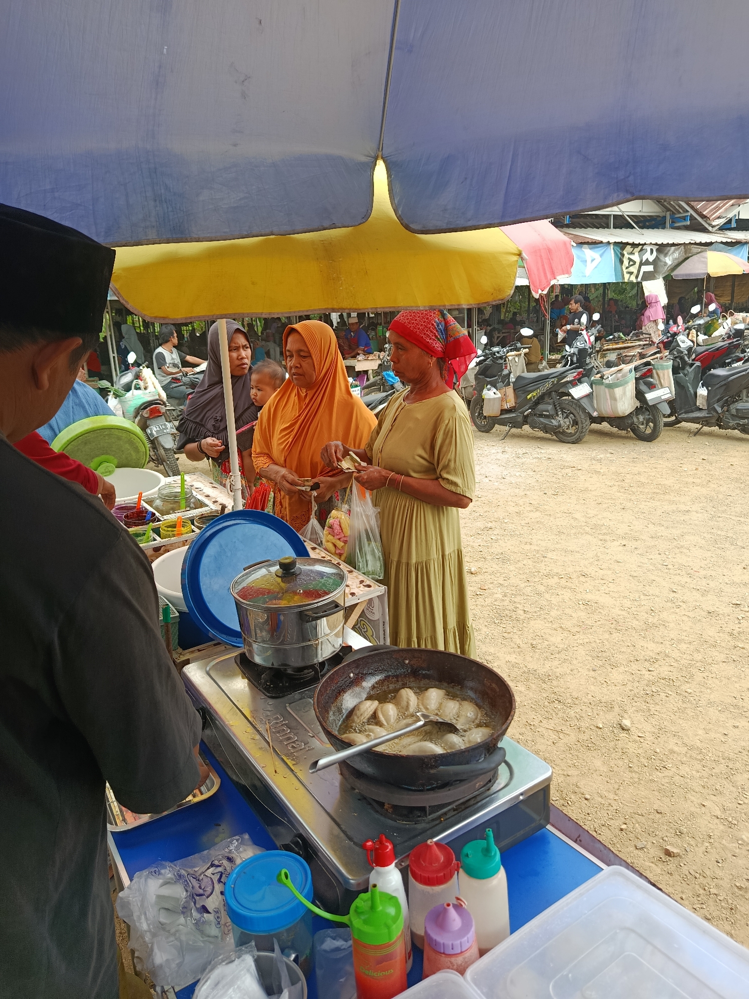 The image depicts an outdoor scene at a market in Sarwadadi, Indonesia, under a large blue and yellow tarp. In the foreground, a person wearing a dark cap and a dark grey shirt is standing on the left, facing away from the camera, partially obscuring a food stall. The stall has a blue tabletop and contains various items including a steamer pot with food inside, colorful bottles of condiments, and plastic containers of food items. To the right of the stall, two women are interacting. One woman, wearing an orange hijab and a light-colored garment, is holding what appears to be money and food packages. The other woman, wearing a red and patterned headscarf and a long, olive-green dress, is looking towards the first woman. Behind them, a child is visible. In the background, numerous motorcycles are parked, and stalls with awnings can be seen, suggesting a bustling market environment. The ground is covered in sand or gravel.