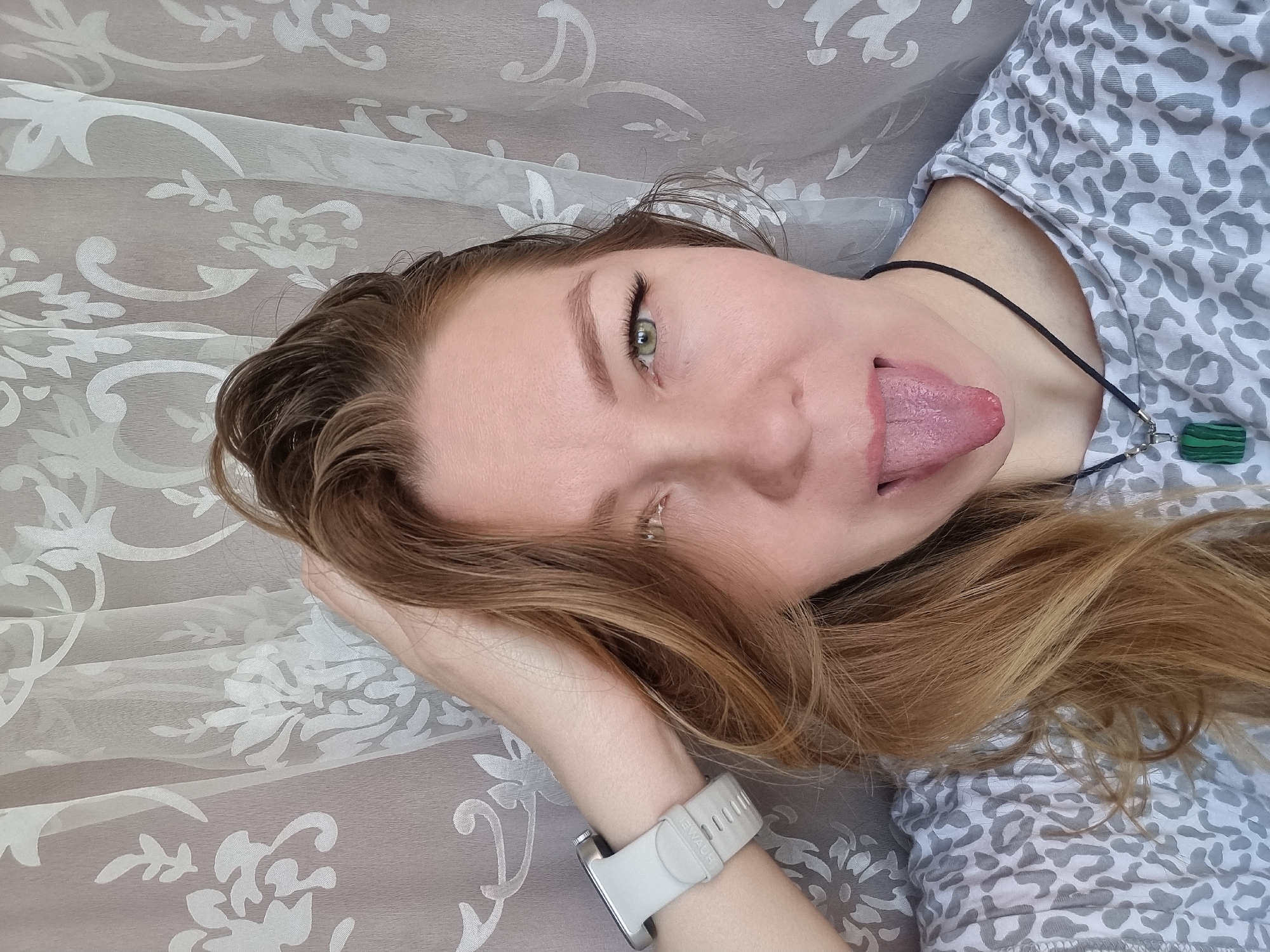 A close-up, high-angle shot captures a person, likely female, lying down with their head tilted to the right, looking directly at the camera. The person has long, light brown hair and green eyes, and is playfully sticking out their tongue. They are wearing a light-colored shirt with a grey and white leopard print pattern. On their left wrist, a light grey smartwatch with the brand name "EWAUH" visible on its band is worn. Around their neck, a black cord necklace features a rectangular pendant made of green and black striped stone, possibly malachite. The background consists of a light-colored fabric, likely a sheet or curtain, adorned with a subtle, raised floral or damask pattern, indicating an indoor setting. The image conveys a casual and lighthearted mood.