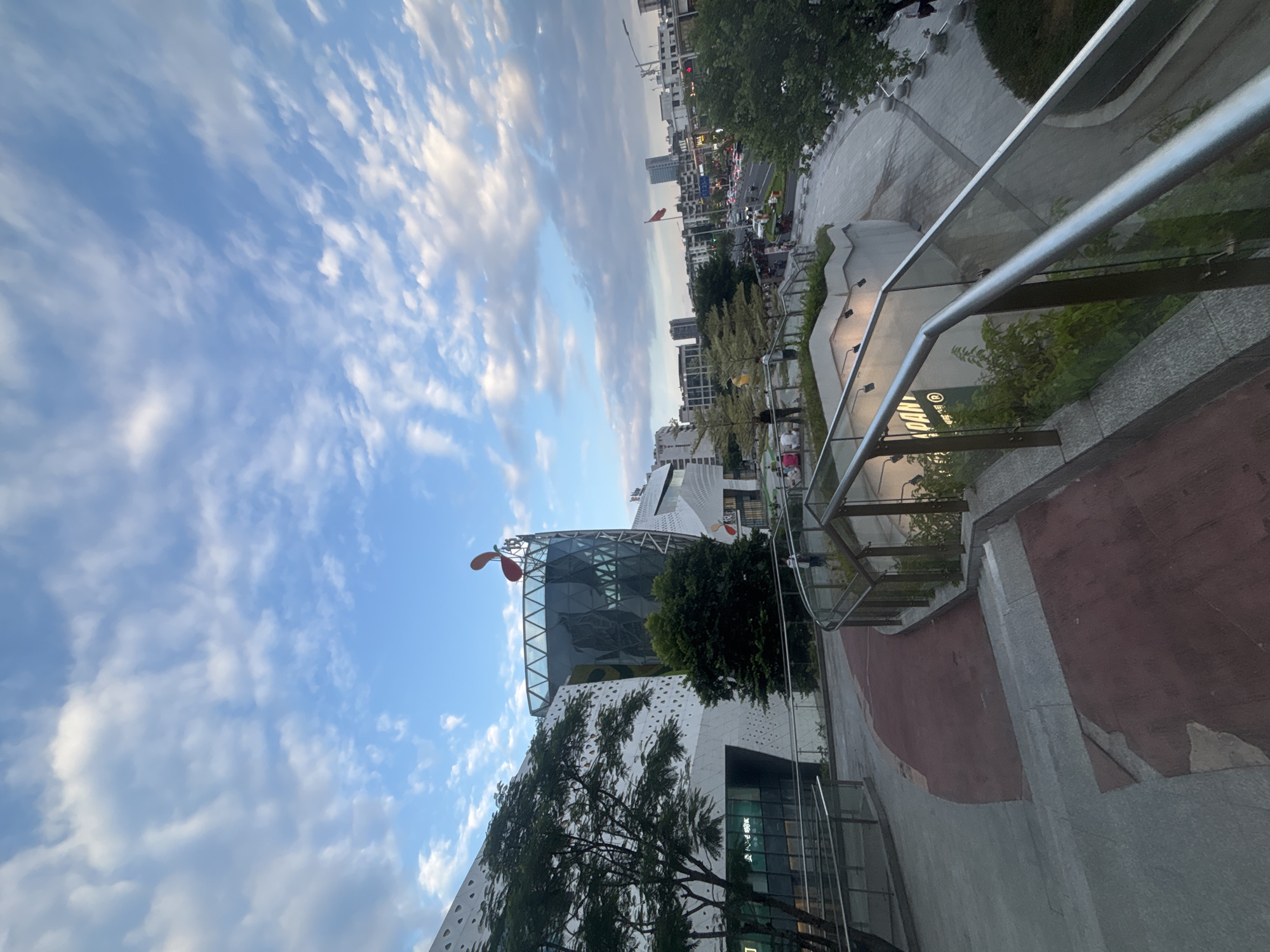 The image depicts an outdoor scene in Liwan District, China, under a sky filled with wispy clouds and patches of blue. In the foreground, a set of stairs with a reddish surface leads down to a paved pathway. A modern building with a white, patterned facade and a glass-enclosed section with a diamond pattern dominates the left side of the frame. A distinctive red, abstract sculpture resembling a flower or a bird's head is visible atop this building. To the right, a curved walkway with glass railings and metal supports guides visitors. Beyond the walkway, trees and other foliage are interspersed. In the background, a street with moderate traffic, including cars and a few bicycles, is visible, lined with multi-story buildings. A Chinese flag flies from a pole in the distance. Several people are scattered throughout the scene, some walking on the pathways, others sitting on benches or near the building. The overall impression is a contemporary urban public space, possibly a commercial or entertainment complex, during daylight hours.