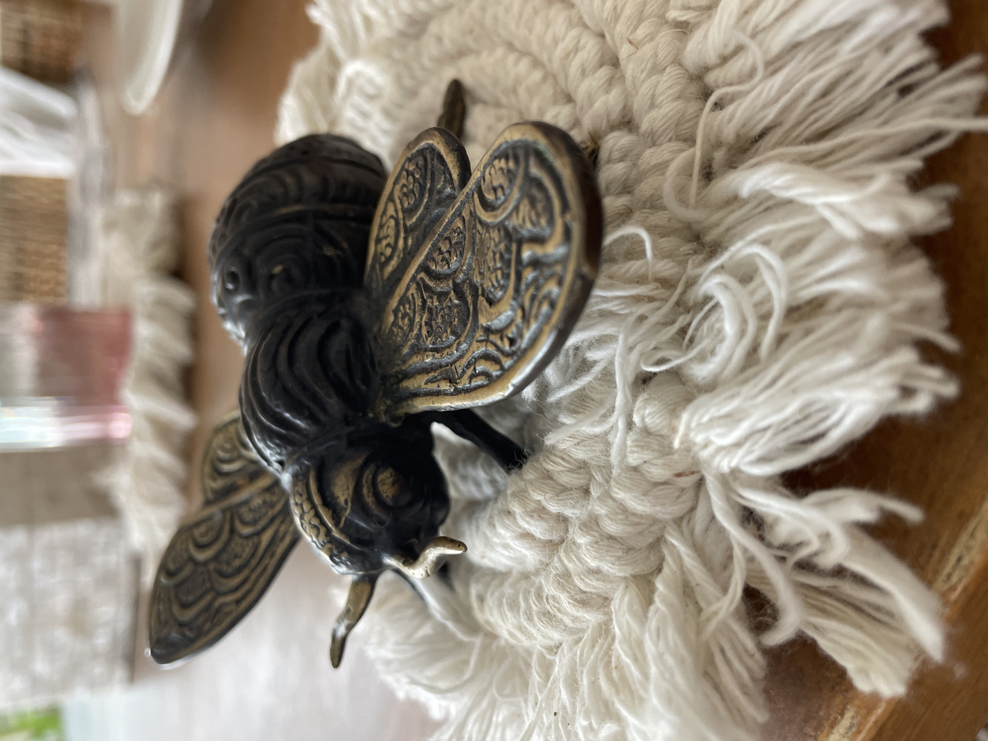 A close-up, high-angle photograph showcases a decorative metallic bee figurine resting on a textured, off-white macrame coaster. The bee, the central subject, is crafted from dark metal, possibly aged brass or bronze, featuring intricate golden-toned patterns on its segmented body and detailed wings, giving it an antique or vintage appearance. Its small legs and antennae are visible, adding to its realistic yet ornamental quality.
The macrame coaster beneath the figurine is a prominent element, characterized by its tightly woven, braided texture in a cream or off-white color, with a soft, feathery fringe extending around its edges. This provides a soft, fibrous contrast to the hard, metallic surface of the bee.
The background is intentionally blurred, indicating a shallow depth of field that keeps the focus sharply on the bee and the coaster. Hints of other objects and surfaces are vaguely discernible in the background, suggesting an indoor setting, possibly a tabletop or shelf. The lighting appears natural and soft, highlighting the metallic sheen of the bee and the delicate textures of the macrame. No specific activity is depicted, as the image focuses on a static decorative display. This scene is set in Kuta Selatan, Indonesia.