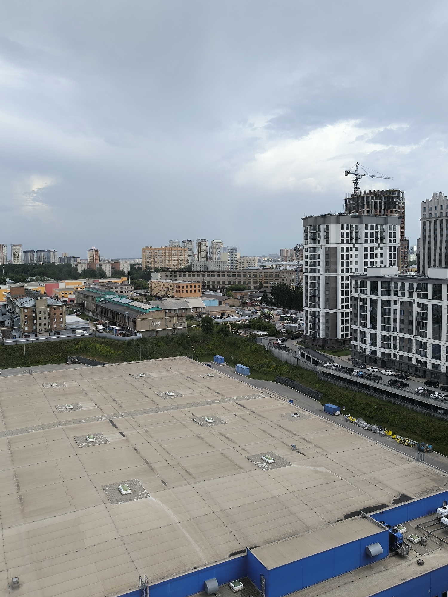 The image depicts a cloudy, overcast day in Novosibirsk, Russia. The foreground is dominated by the flat, concrete roof of a large, blue-sided building, possibly a warehouse or industrial facility, with numerous square vents and skylights. In the middle ground, a steep, green, grassy embankment slopes down towards a road where cars are parked. Beyond the embankment, a multi-story parking garage filled with vehicles is visible. Further into the distance, a collection of buildings of varying sizes and architectural styles form the city skyline. Several low-rise industrial or commercial buildings with brick and concrete facades are present, interspersed with taller, modern apartment buildings. A construction crane hovers over a tall building under construction, indicating ongoing development in the area. The overall impression is of an urban environment with a mix of older industrial structures and newer residential and commercial developments under a gloomy sky.