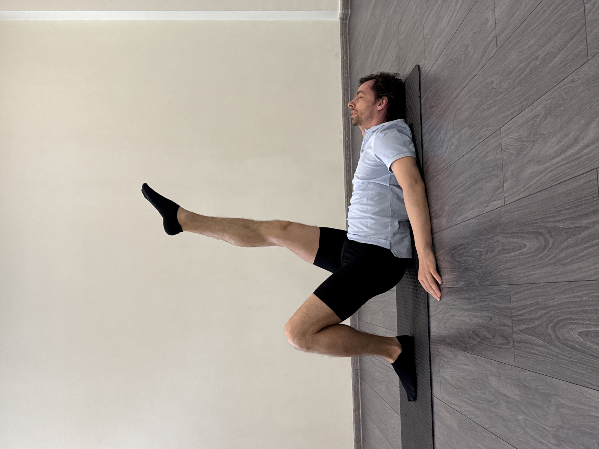 A man is lying on his back on a yoga mat against a gray wall. He is wearing black shorts, a light blue polo shirt, and black socks. His legs are bent, with one leg extended straight up in the air. The yoga mat is dark gray and rectangular. The wall to the left is off-white. The floor is a gray wood-look laminate. The location is Unknown, Unknown.