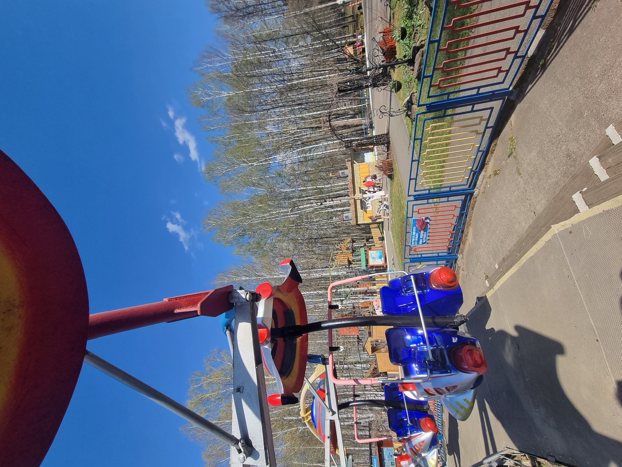 The image captures an outdoor scene in Unknown, Unknown. A children's amusement park ride, designed with blue and red airplane-shaped seats, dominates the foreground. The ride is attached to a red central pole. The background features a grove of tall, slender trees with light green leaves, a blue sky with scattered clouds, a colorful fence, and a paved pathway. The scene seems devoid of people at the moment the picture was taken.